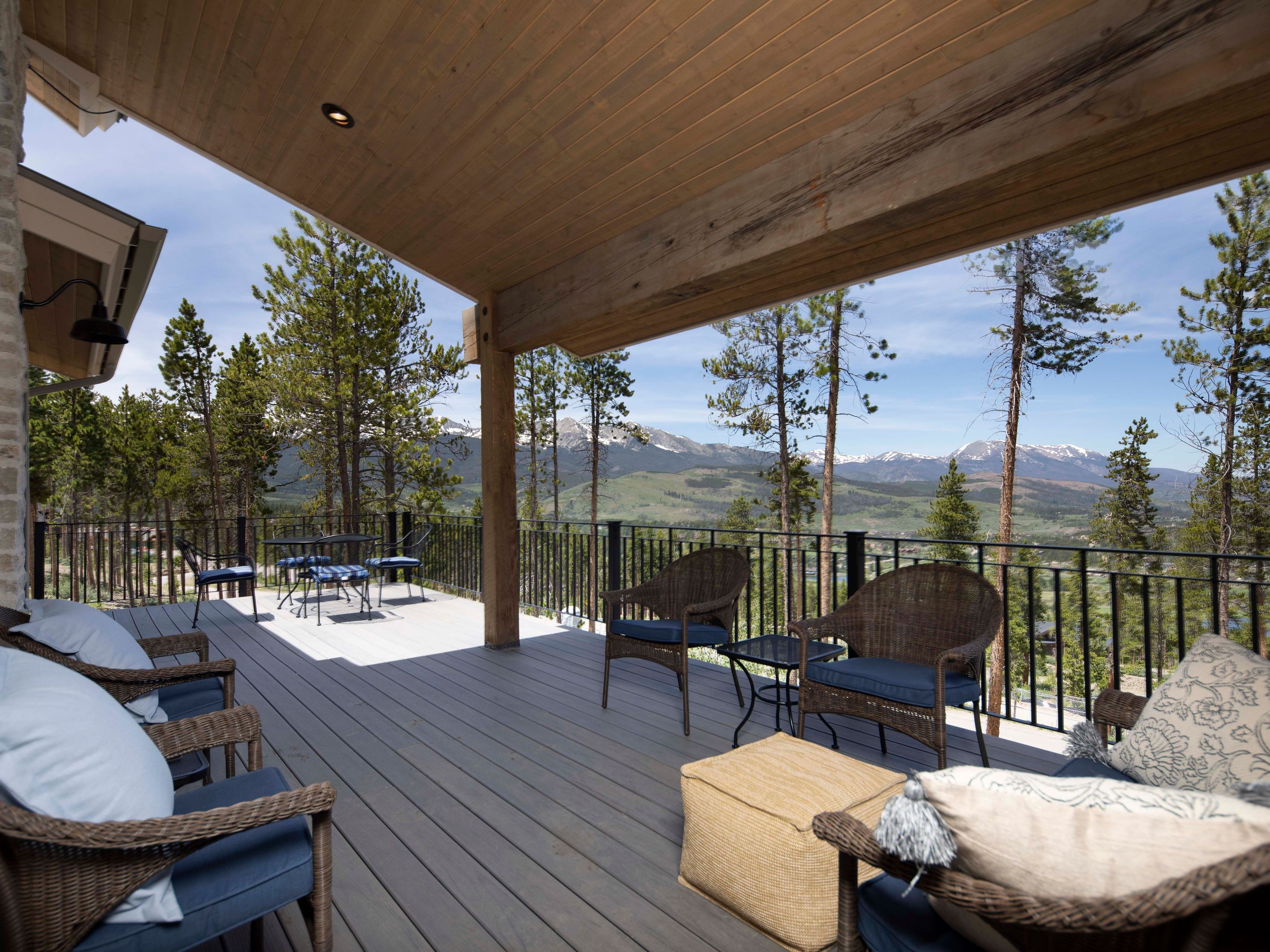 This expansive covered deck offers a serene outdoor living space with comfortable wicker seating and a neutral-toned ottoman. The area features a dark metal railing that frames a breathtaking, panoramic view of rolling green hills and snow-capped mountains in the distance. The warm wood-paneled ceiling and sturdy timber support beam create a rustic, inviting atmosphere perfect for enjoying the mountain scenery.