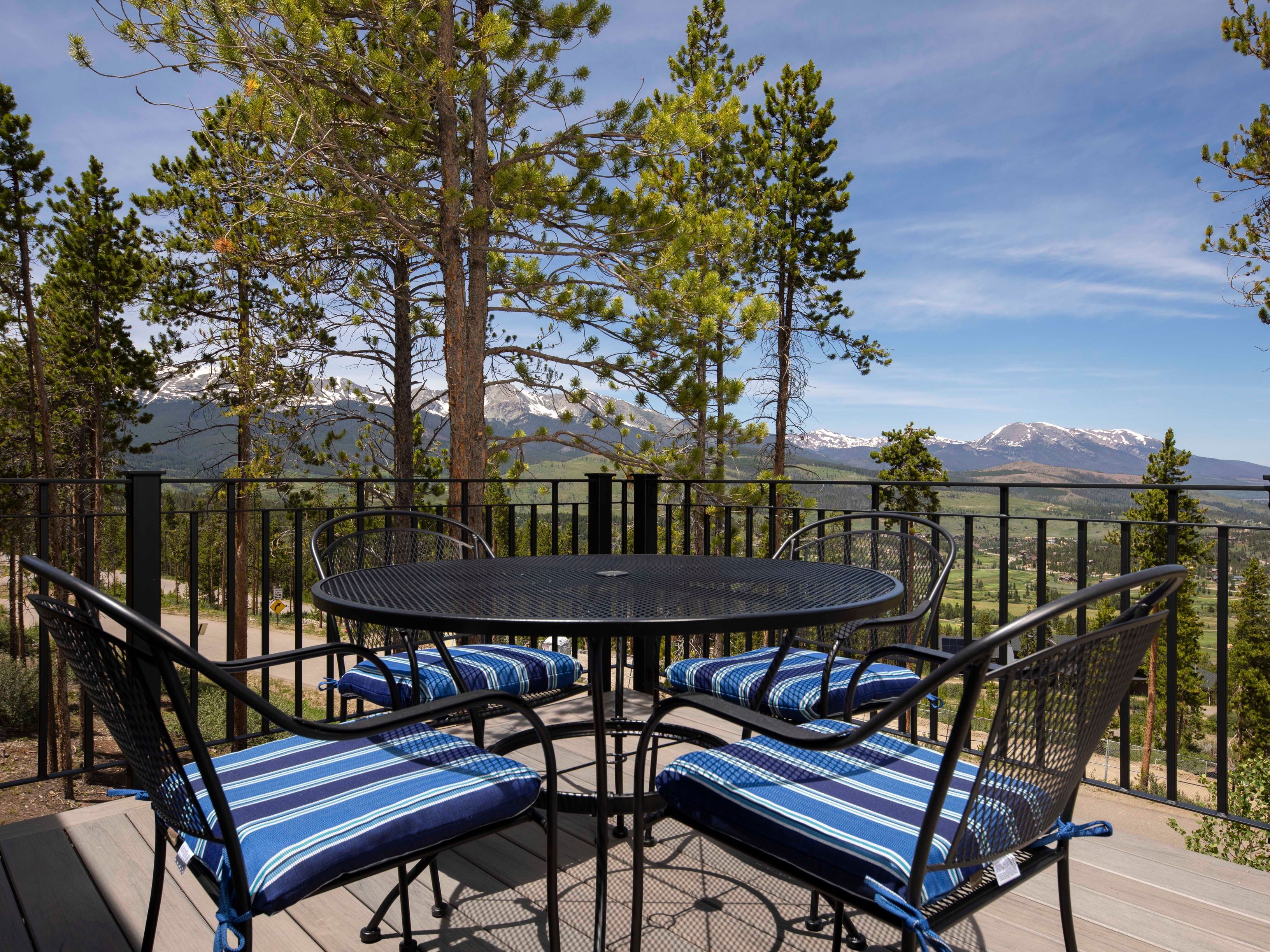 This elevated deck features a black metal dining set with blue and white striped cushions, perfectly positioned to take in the expansive mountain views. The scene is framed by tall pine trees and a black metal railing, creating a serene outdoor living space. The perspective is eye-level, capturing the inviting atmosphere of the deck against a backdrop of snow-capped peaks and a clear blue sky.
