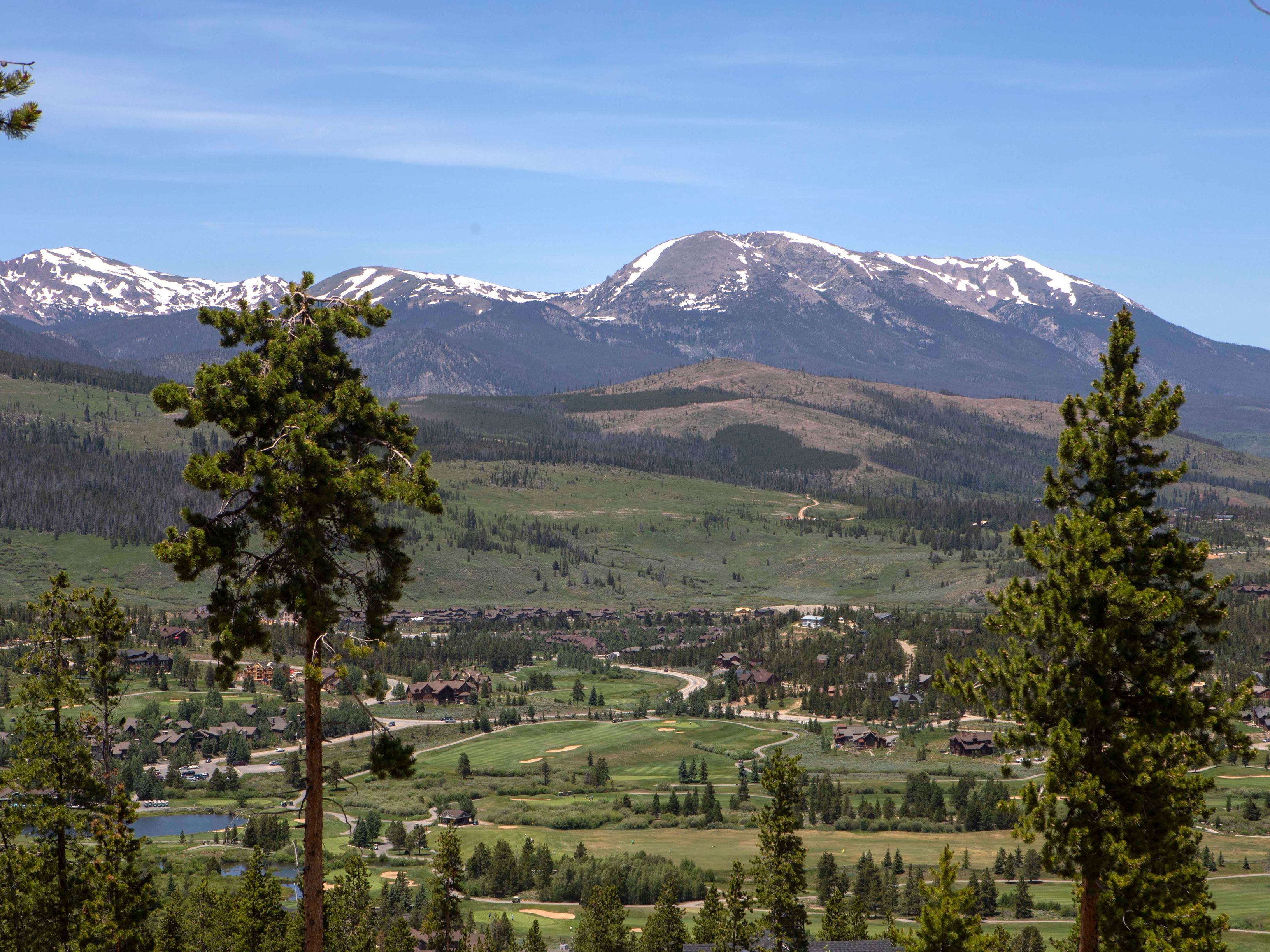 This expansive aerial view captures a picturesque mountain valley community nestled at the base of majestic, snow-capped peaks. The landscape features a lush green golf course winding through a residential neighborhood of homes with rustic, wood-toned architecture. Two prominent pine trees frame the foreground, providing a natural depth that draws the eye toward the serene valley floor under a clear blue sky.