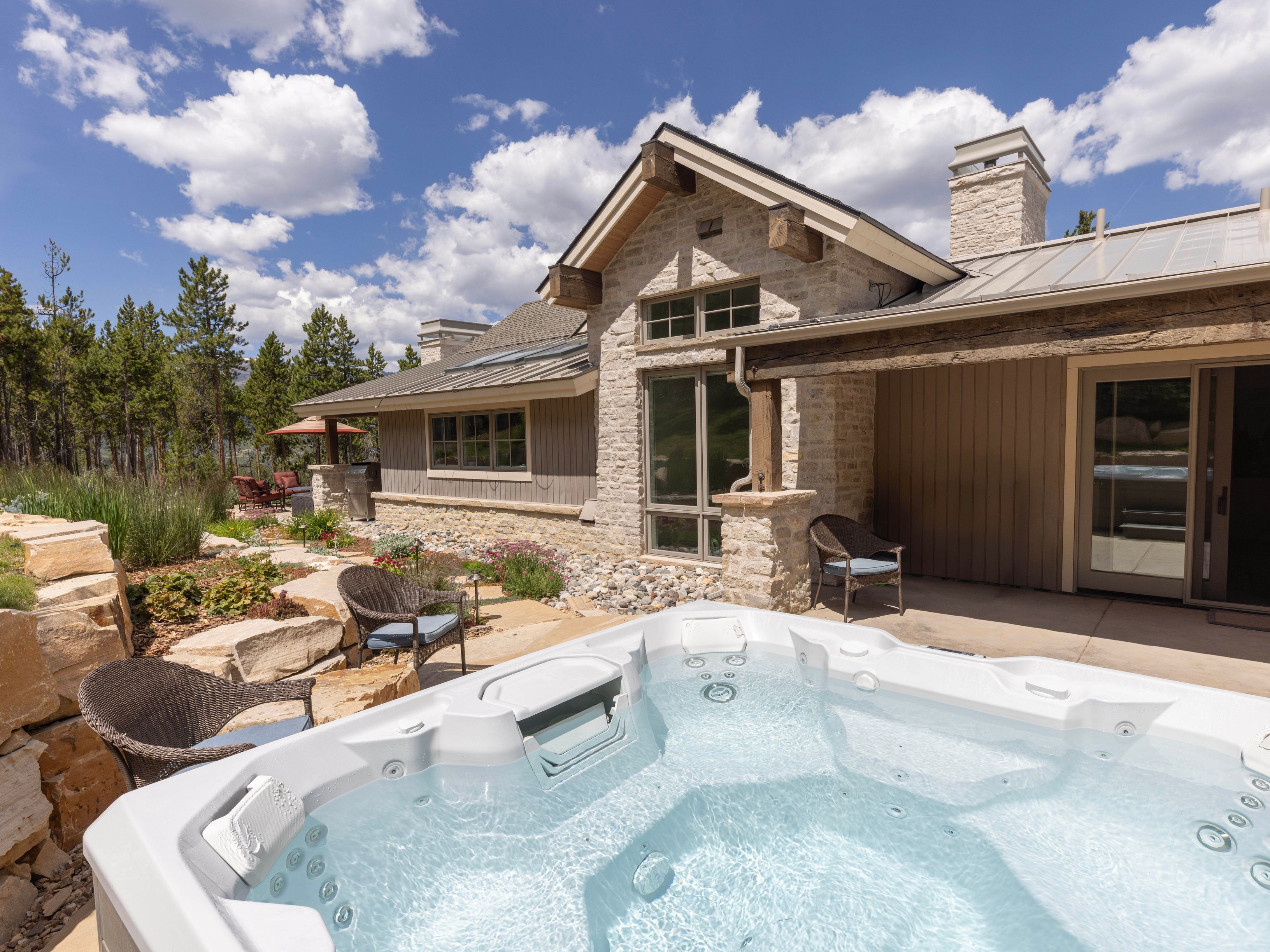 This outdoor living space features a large, modern hot tub in the foreground, set against a rustic-style home with stone and wood siding. A covered patio area provides a shaded seating spot with wicker chairs, while the surrounding landscape includes natural stone retaining walls and native plantings. The scene is captured from a slightly elevated perspective, showcasing the seamless transition between the home's architecture and the mountainous, wooded environment.
