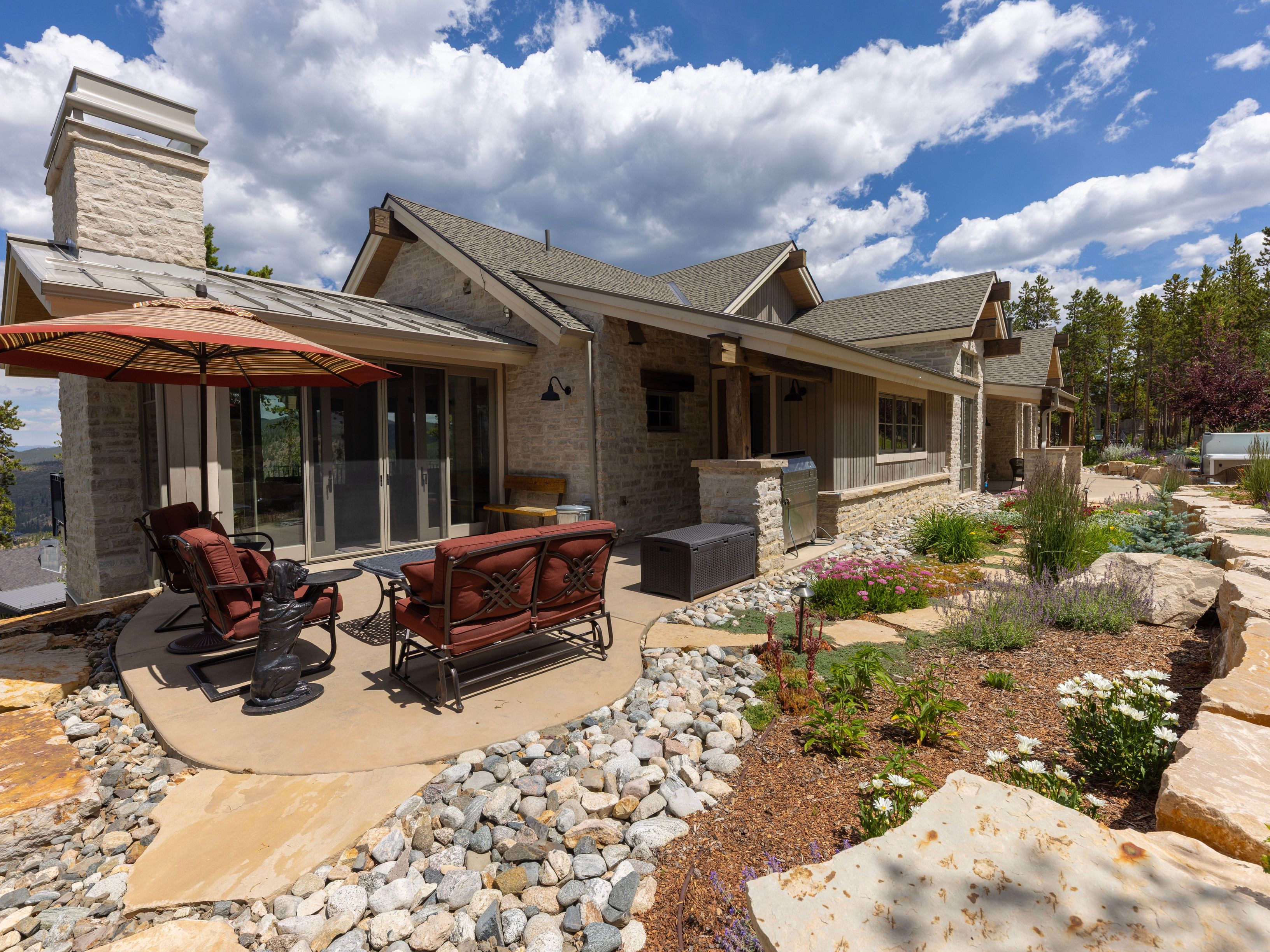 This inviting outdoor living space features a concrete patio furnished with a comfortable seating area, including a sofa and chairs shaded by a large umbrella. The home showcases a rustic aesthetic with stone-clad walls, a prominent stone chimney breast, and a metal roof. A landscaped garden with native plants, decorative rocks, and large stone pavers leads the eye toward the surrounding forest, creating a serene and picturesque mountain retreat.
