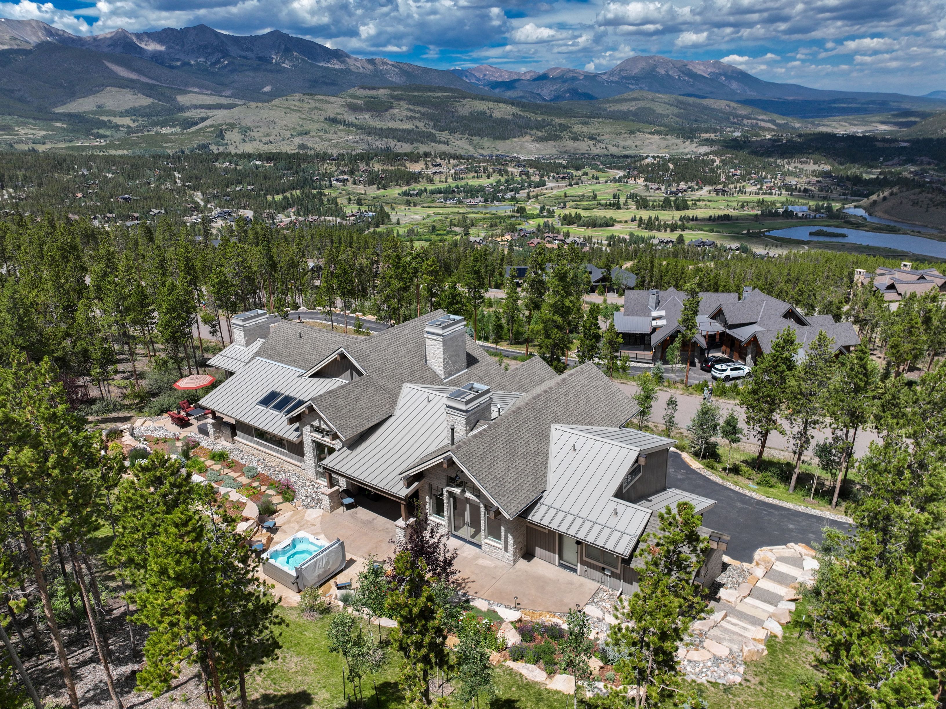 This high-angle aerial view captures a sprawling, modern mountain residence nestled within a dense pine forest. The home features a complex roofline with metal and shingle sections, multiple stone chimneys, and a spacious patio area complete with a hot tub. In the background, a scenic valley with a golf course and a lake unfolds beneath a dramatic mountain range under a bright, cloudy sky.
