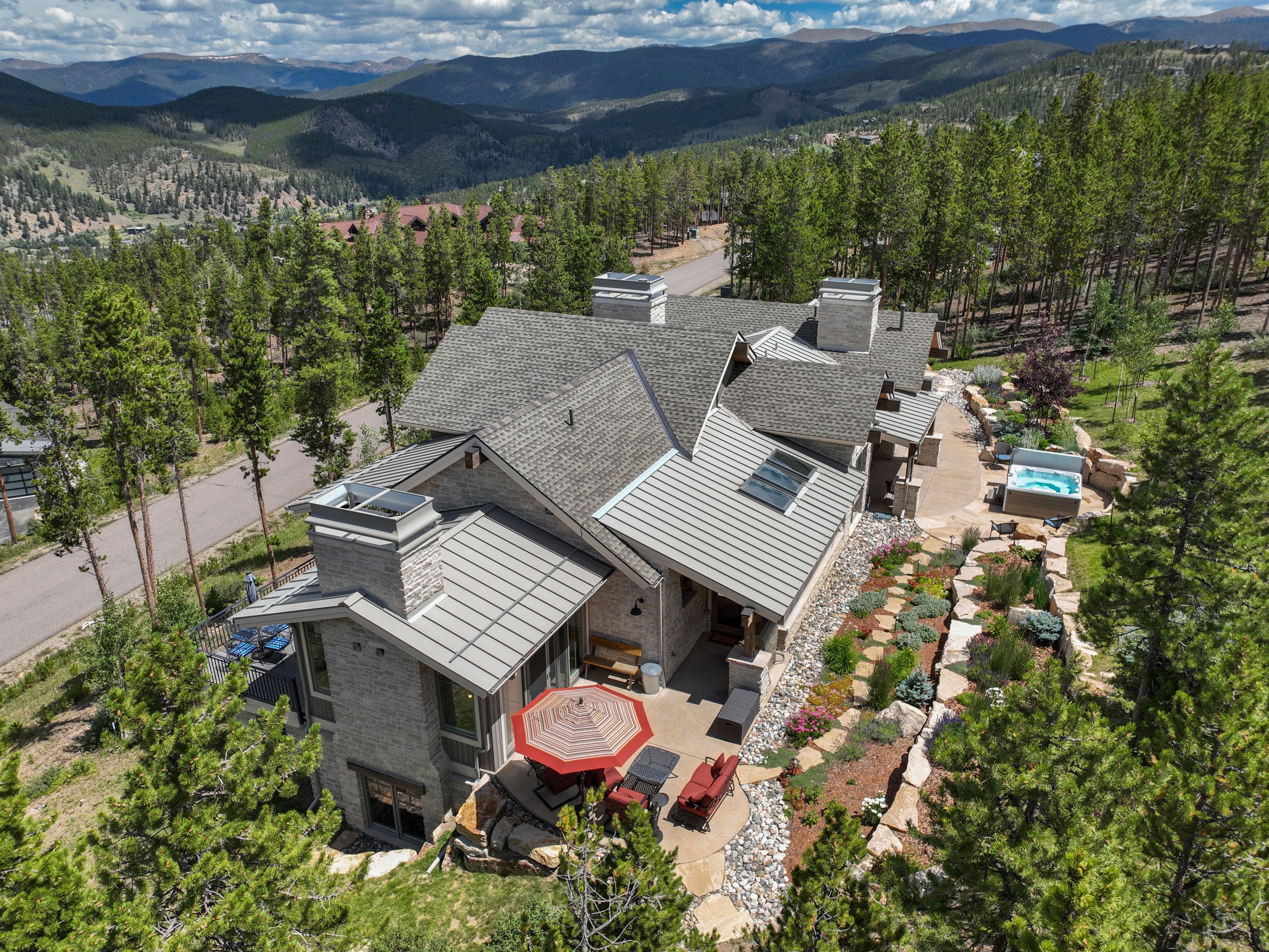 This high-angle aerial perspective captures a luxurious mountain residence nestled within a dense pine forest. The home features a multi-gabled roofline with metal and shingle sections, a stone-clad exterior, and a spacious rear patio complete with a dining set and a hot tub. The surrounding landscape is characterized by tiered stone retaining walls, natural rock gardens, and sweeping views of distant mountain ranges under a bright, cloudy sky.