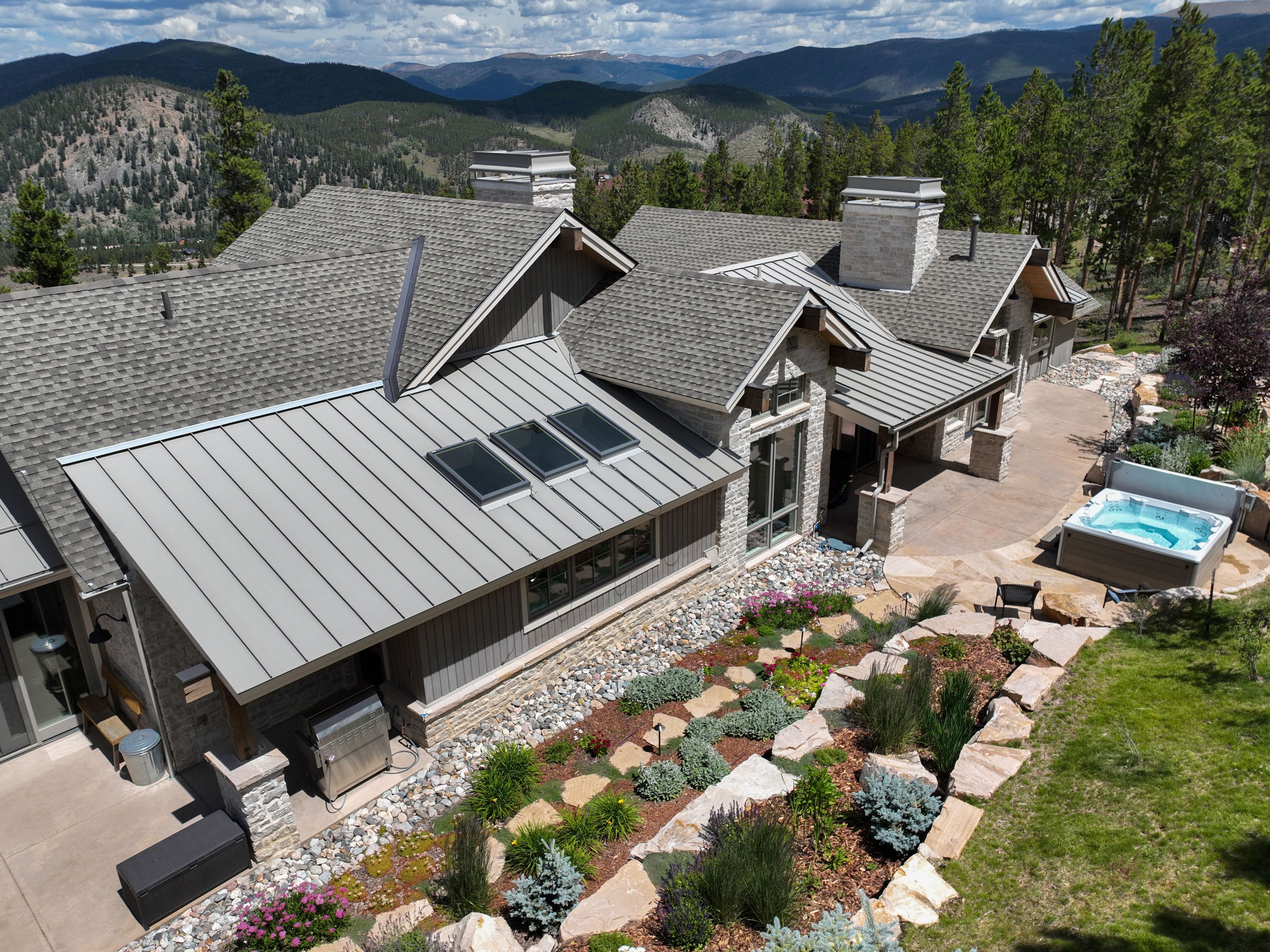 This high-angle aerial view captures a luxurious mountain residence featuring a blend of stone and metal roofing, set against a backdrop of rolling forested hills and distant peaks. The property includes a landscaped terraced garden with stone retaining walls, a private hot tub on a concrete patio, and a stainless steel outdoor grill area. The architecture emphasizes a rustic yet modern aesthetic, perfectly integrated into the rugged natural landscape.