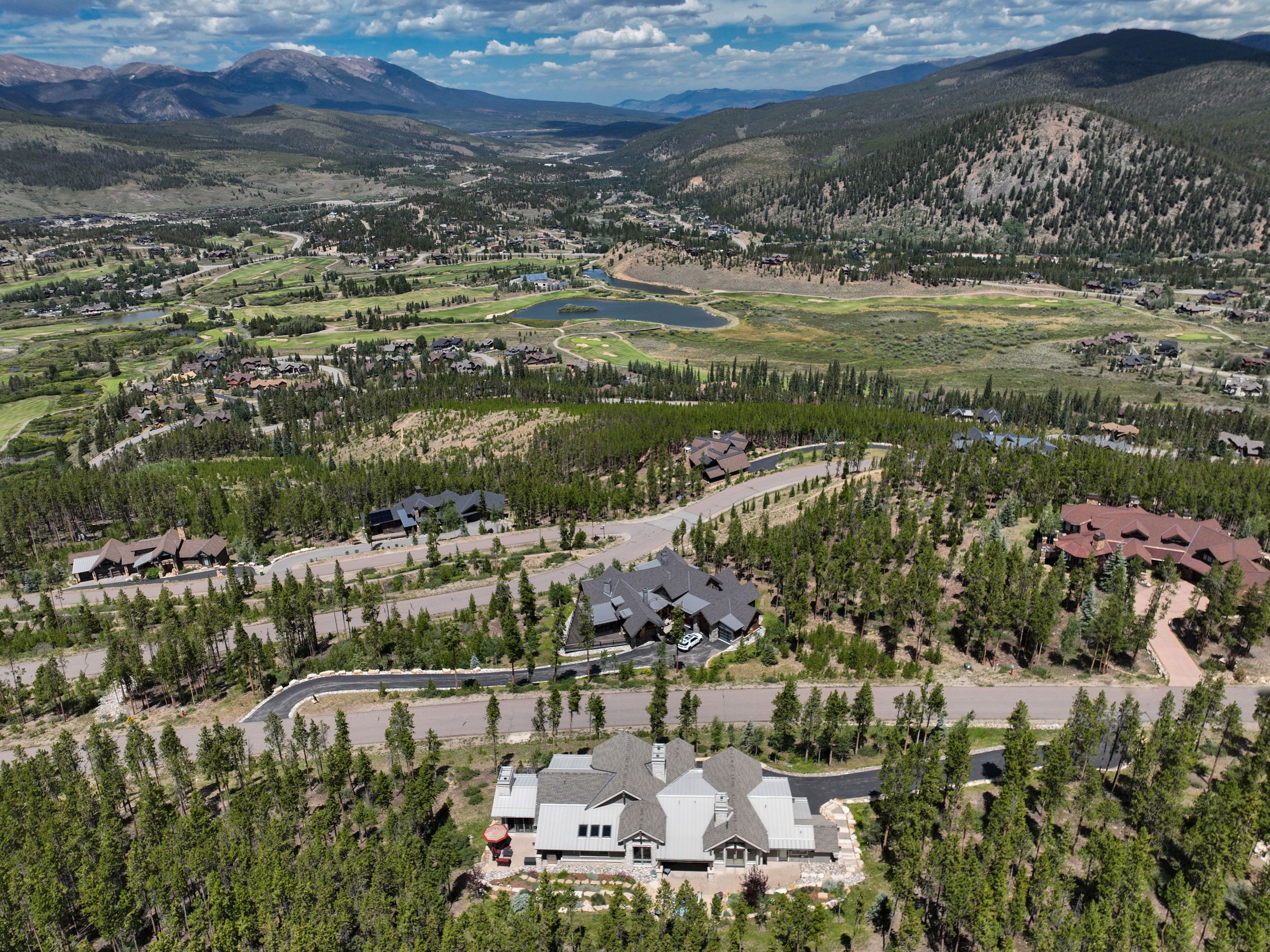 This high-angle aerial perspective showcases a sprawling mountain estate nestled within a lush, forested landscape. The property features a large, multi-gabled residence with a light-colored roof, surrounded by mature pine trees and winding paved roads. In the distance, a scenic valley unfolds with a golf course, a small lake, and majestic mountain ranges under a bright, cloud-dappled sky.