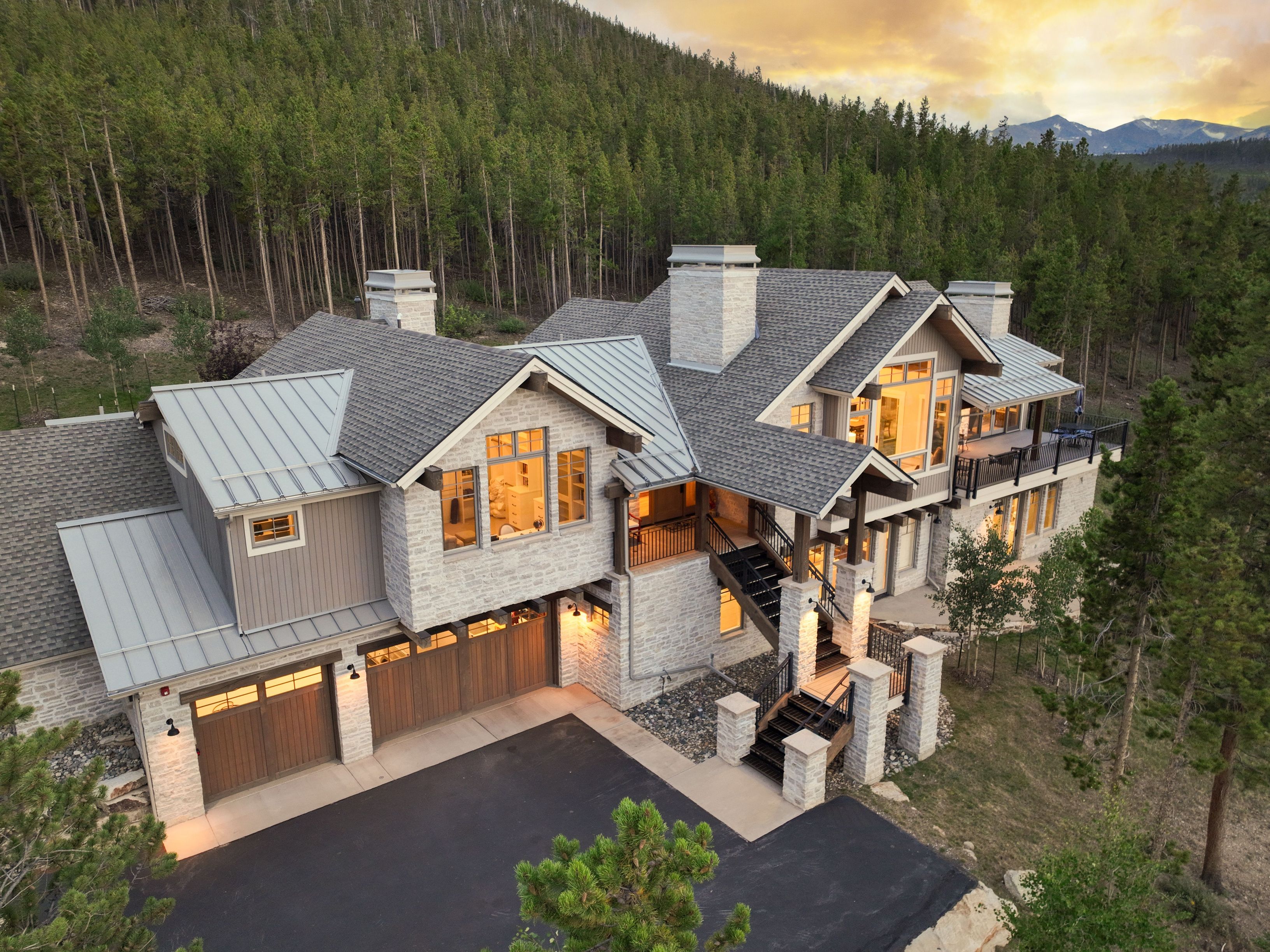 This elevated aerial perspective captures a sprawling mountain estate nestled against a dense forest backdrop during a golden sunset. The home features a sophisticated blend of light-toned stone masonry, vertical wood siding, and multi-gabled rooflines with metal and shingle accents. A paved driveway leads to a multi-car garage, while an exterior staircase provides access to the upper levels, highlighting the home's integration into the rugged, sloped landscape.