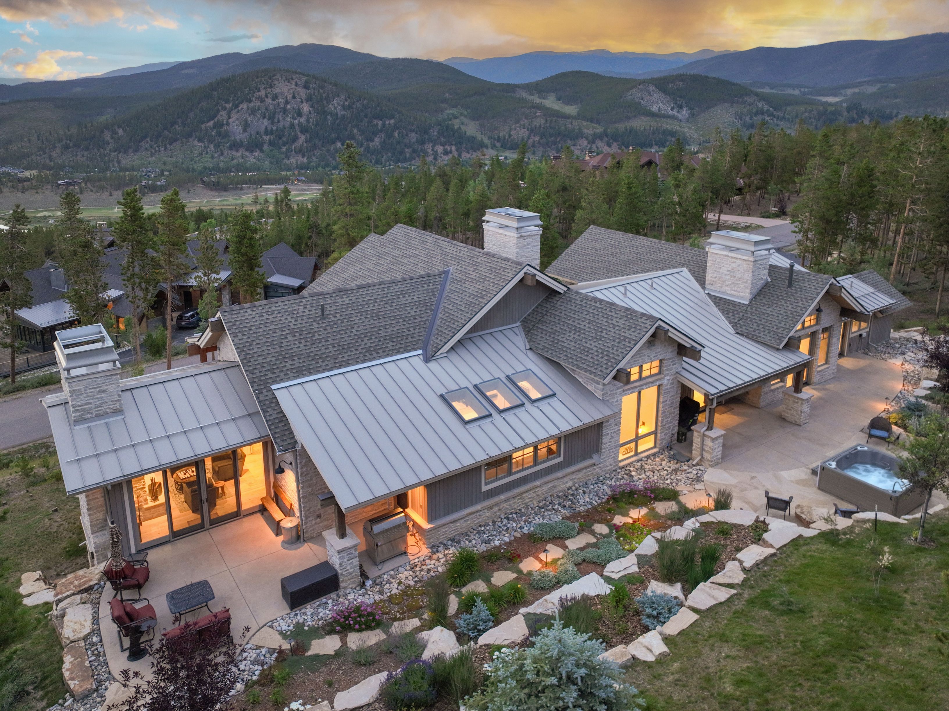 This elevated aerial perspective captures a luxurious mountain residence at dusk, showcasing a sophisticated blend of stone and dark siding materials. The home features multiple metal-roofed sections, skylights, and expansive patios that integrate seamlessly with the rugged, landscaped terrain and a private hot tub. The backdrop of rolling mountains under a vibrant sunset sky creates a serene and high-end atmosphere.