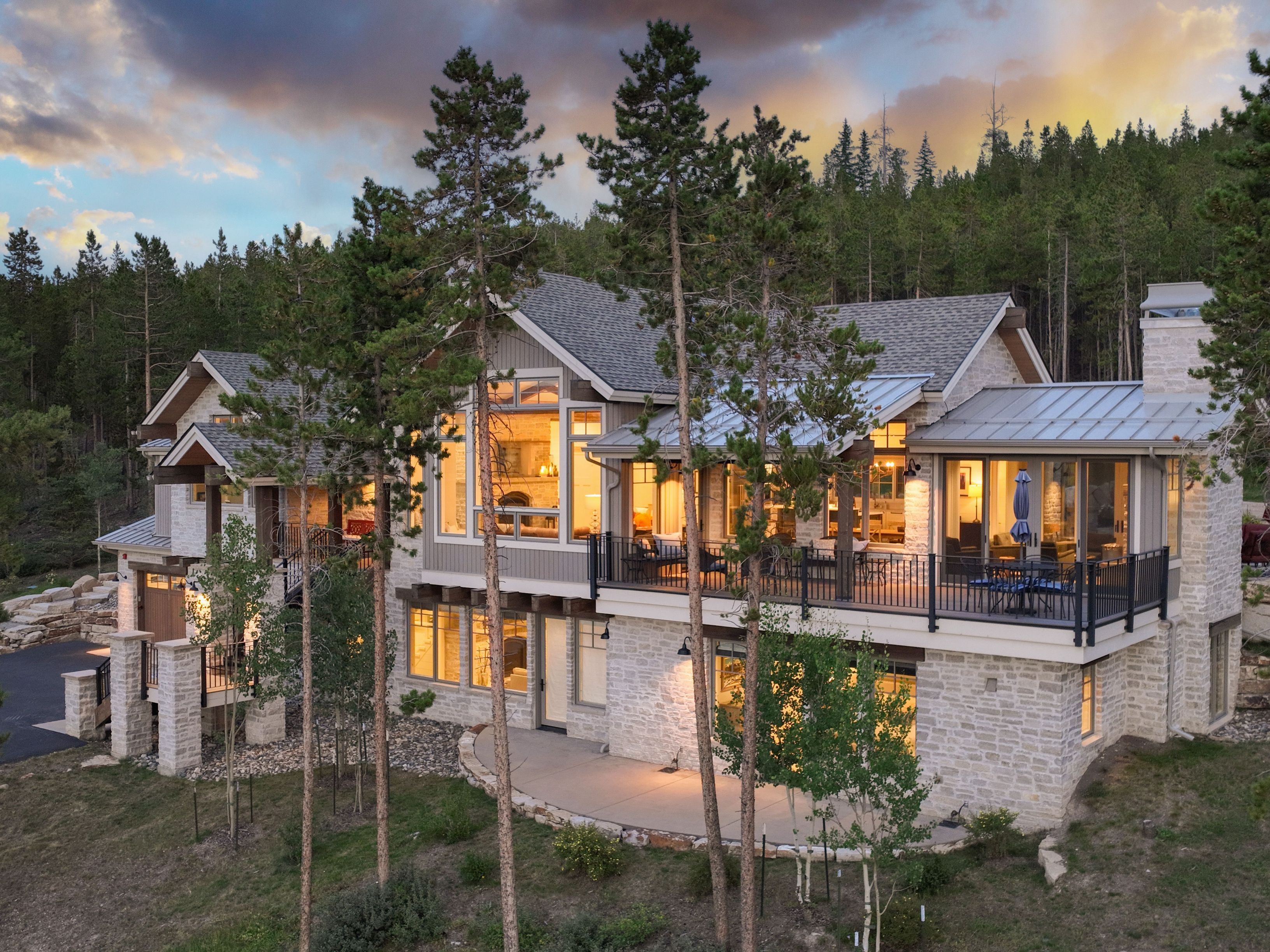 This stunning mountain residence features a sophisticated blend of light-toned stone masonry and warm-toned wood siding, set against a backdrop of dense pine forest. The home showcases a multi-level design with a spacious elevated deck, large windows that capture the sunset light, and a lower-level walk-out patio. The architectural style is a refined take on modern rustic, emphasizing seamless integration with the natural, sloped landscape.