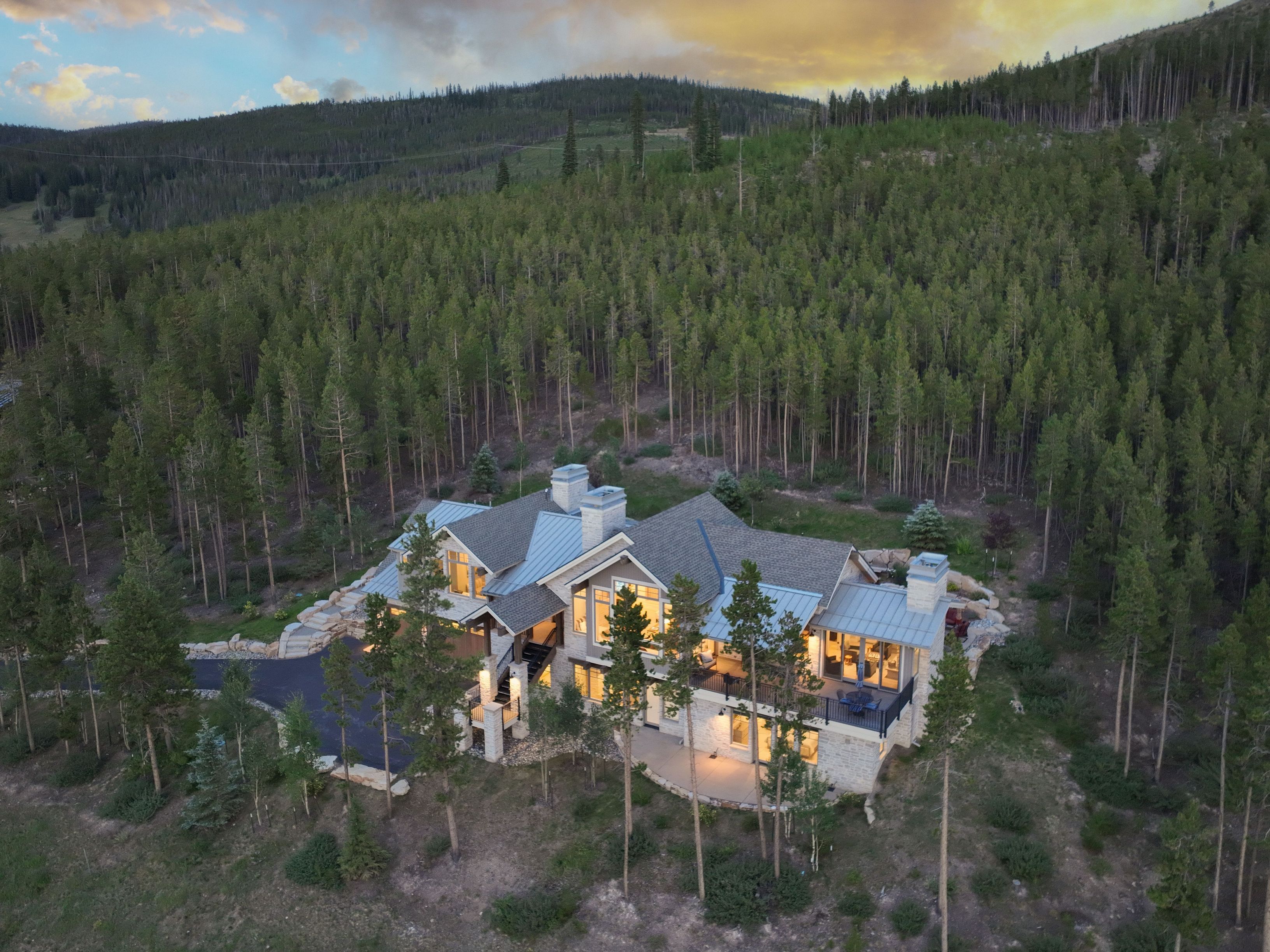 This aerial perspective captures a luxurious mountain residence nestled into a dense, evergreen-forested hillside at dusk. The home features a multi-gabled roofline with metal accents, stone exterior walls, and expansive windows that glow warmly from within. A paved driveway leads to the entrance, while a tiered stone patio and elevated deck provide seamless transitions between the indoor living spaces and the rugged natural landscape.
