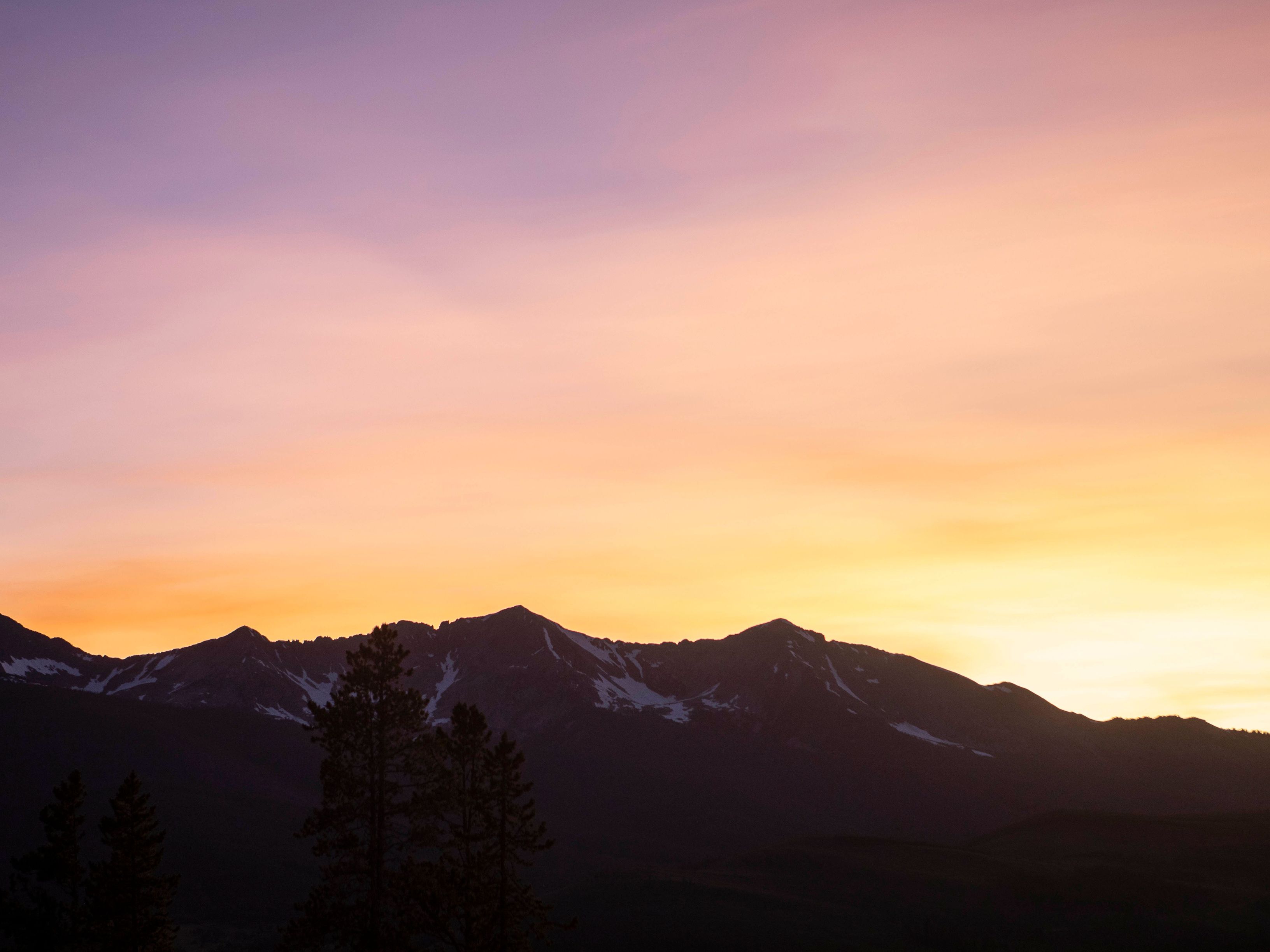 This scenic landscape shot captures a dramatic mountain range silhouetted against a vibrant, multi-colored sunset sky. In the foreground, the dark outlines of evergreen trees frame the lower portion of the view, emphasizing the scale of the peaks. The image evokes a sense of tranquility and natural beauty, highlighting the desirable outdoor surroundings of the property.
