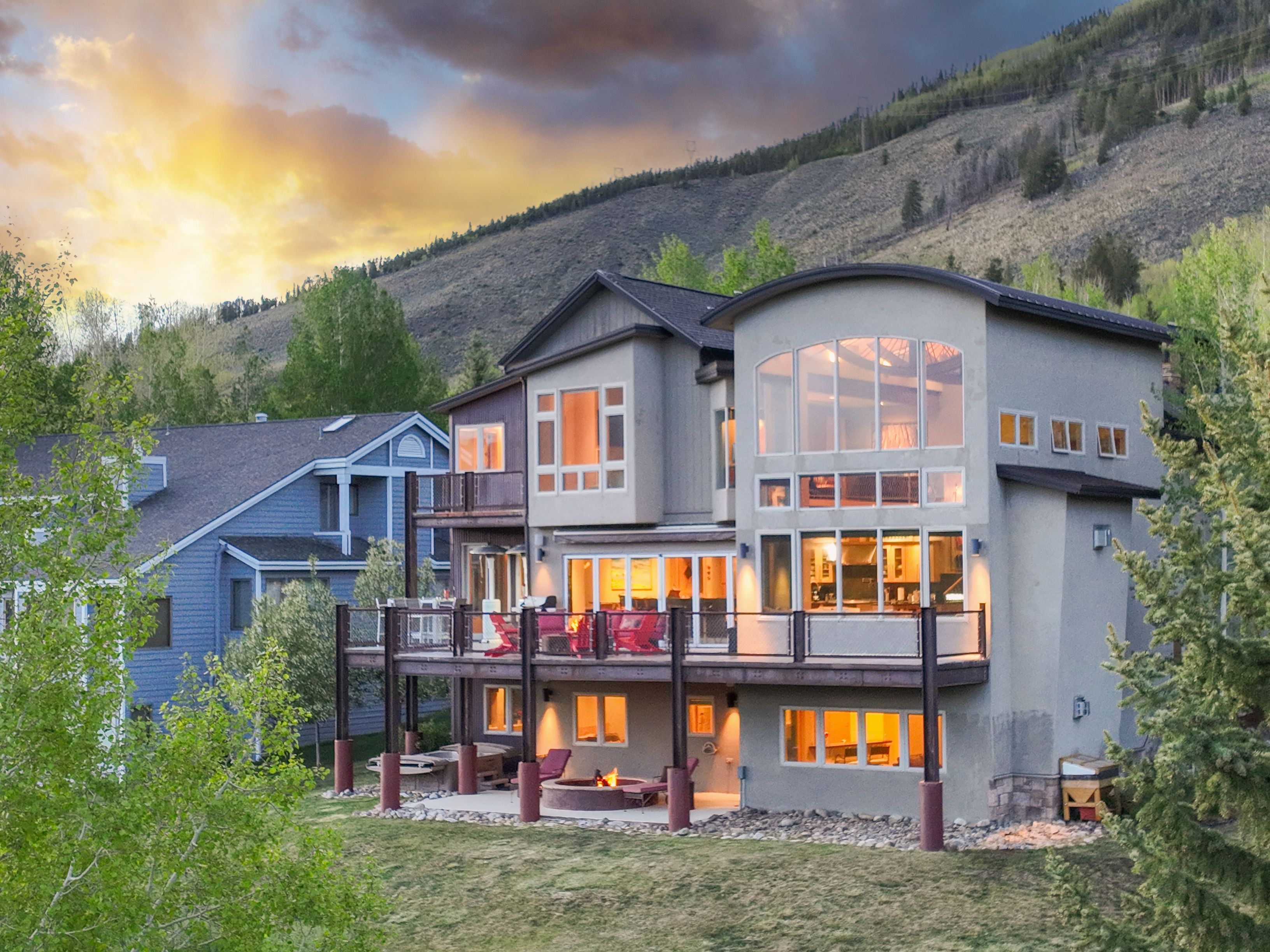 This multi-level mountain home features a contemporary design with a mix of siding textures and a prominent curved roofline. A spacious elevated deck with red seating overlooks a lower patio area equipped with a circular fire pit, all set against a backdrop of a scenic hillside at sunset. The warm interior lighting glows through the large windows, creating an inviting atmosphere for this expansive residential property.