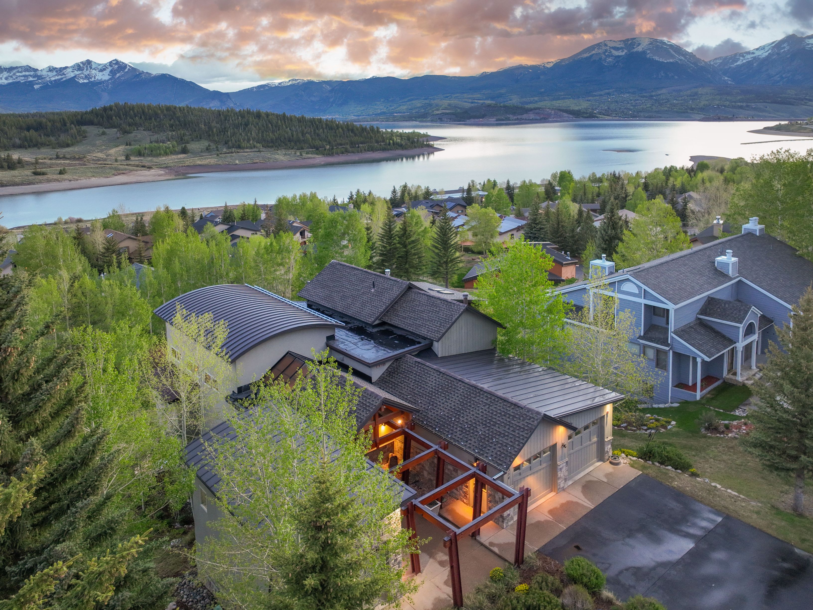 This high-angle aerial shot captures a contemporary mountain home nestled among lush green trees, featuring a unique curved metal roof and a prominent timber-framed entryway. The property is situated in a scenic landscape with a large lake in the background and snow-capped mountains under a dramatic sunset sky. The composition highlights the home's integration into its natural surroundings, showcasing its architectural character and prime location.