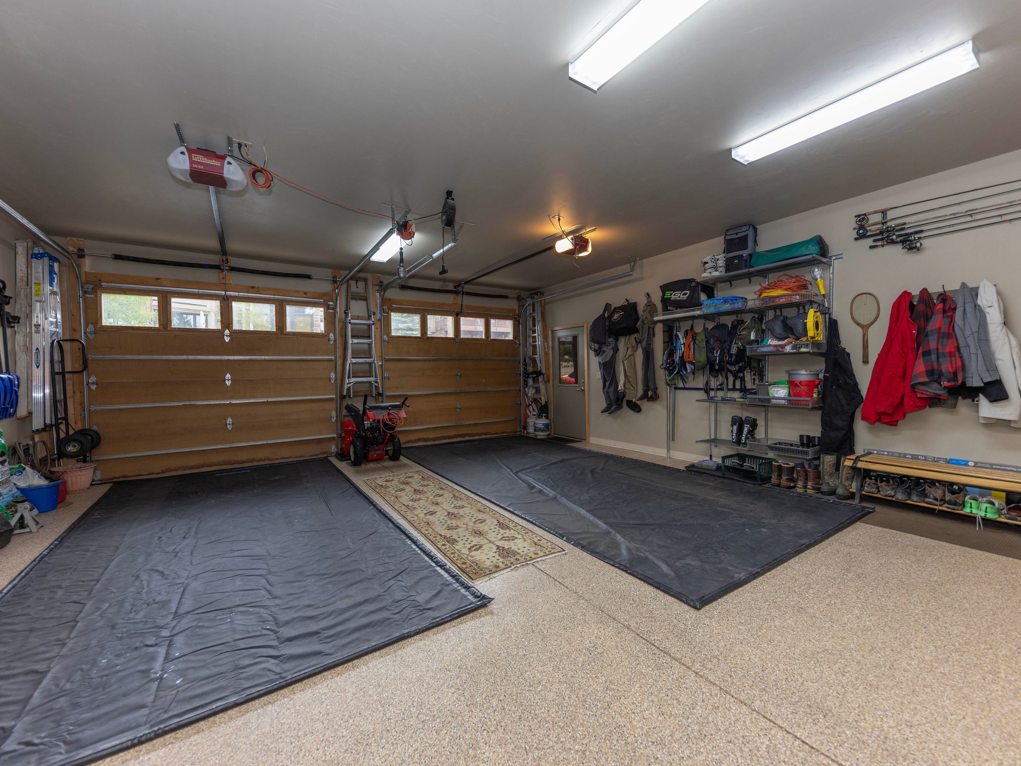 This spacious, well-organized two-car garage features a durable epoxy-coated floor with protective black mats laid out for vehicle parking. The space is equipped with ample storage, including metal shelving units, wall-mounted hooks for outdoor gear, and a bench for footwear. Bright overhead fluorescent lighting illuminates the area, which also includes a small snowblower and a decorative runner rug between the parking bays.