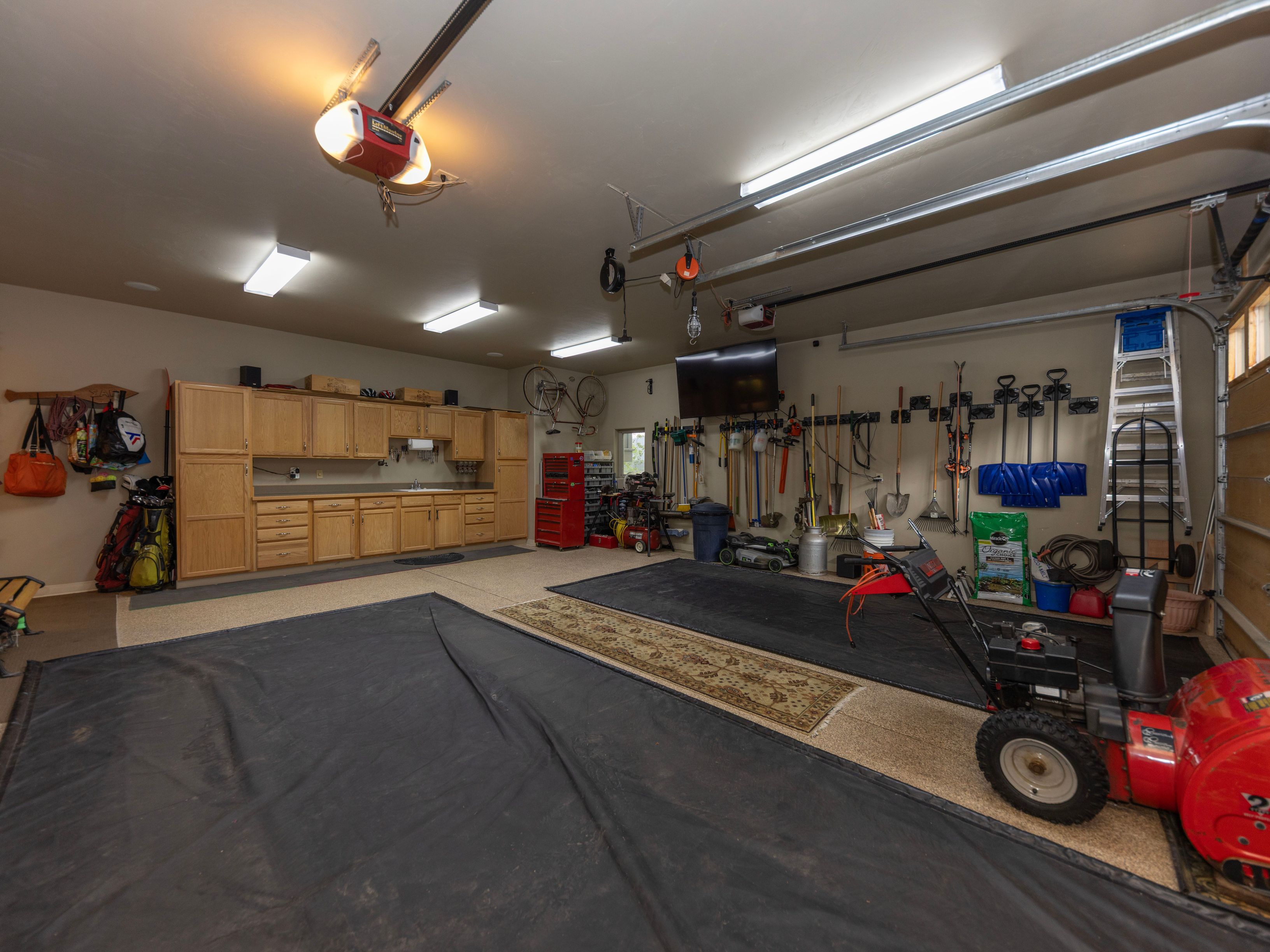 This spacious, well-organized garage features light wood cabinetry with ample counter space and a sink, providing a functional workspace. The area is equipped with overhead lighting, a wall-mounted television, and extensive wall-mounted storage for tools, sports equipment, and a ladder. A large red snowblower sits in the foreground, while protective floor mats cover the epoxy-coated concrete floor, creating a clean and versatile environment.