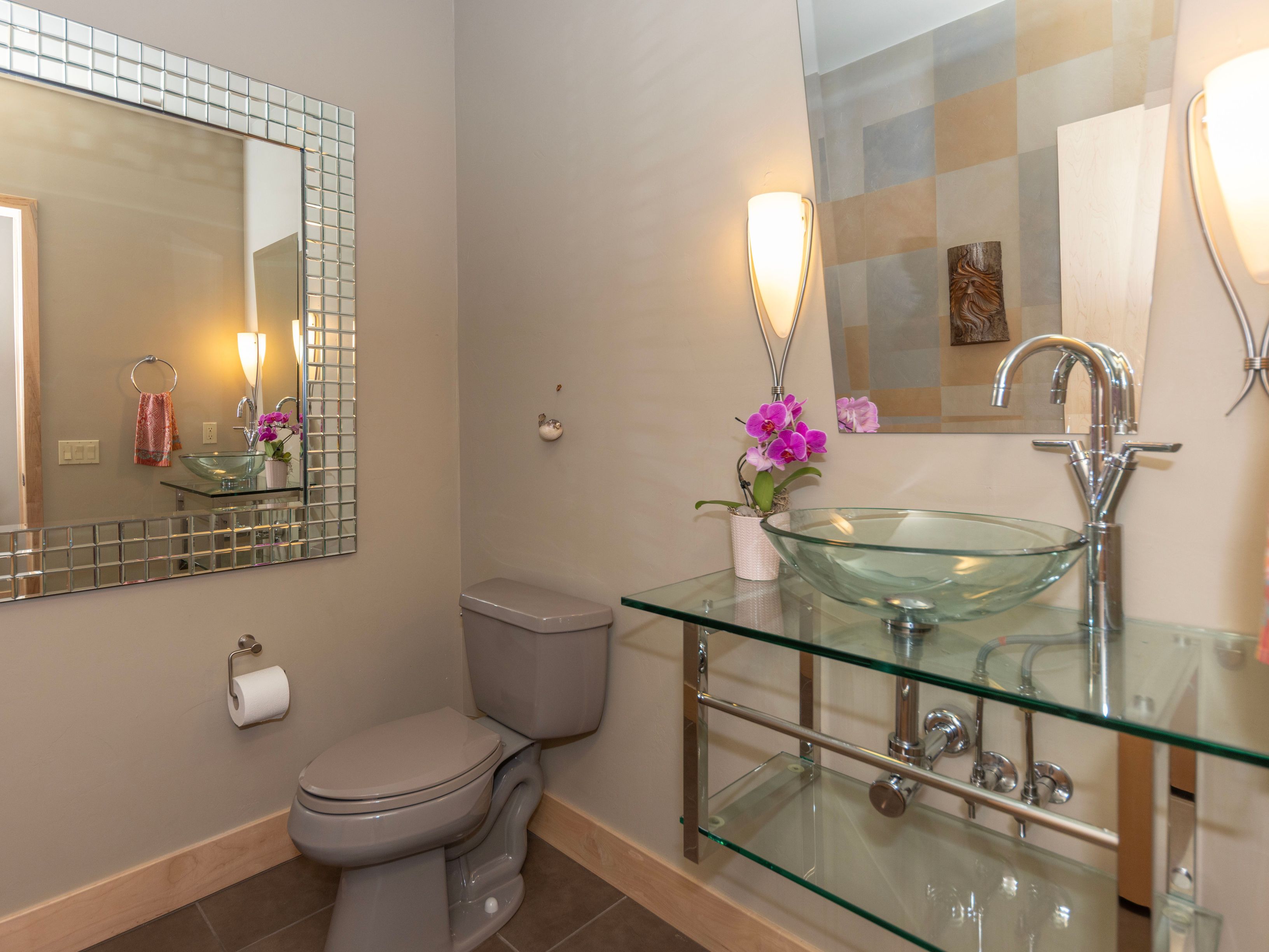 This contemporary powder room features a unique glass vessel sink mounted on a glass vanity with a chrome frame. A large, square-tiled mirror adds a decorative touch, complemented by a second mirror above the sink and warm wall sconces. The space is finished with a neutral-toned toilet and a small potted orchid, creating a clean and modern aesthetic.