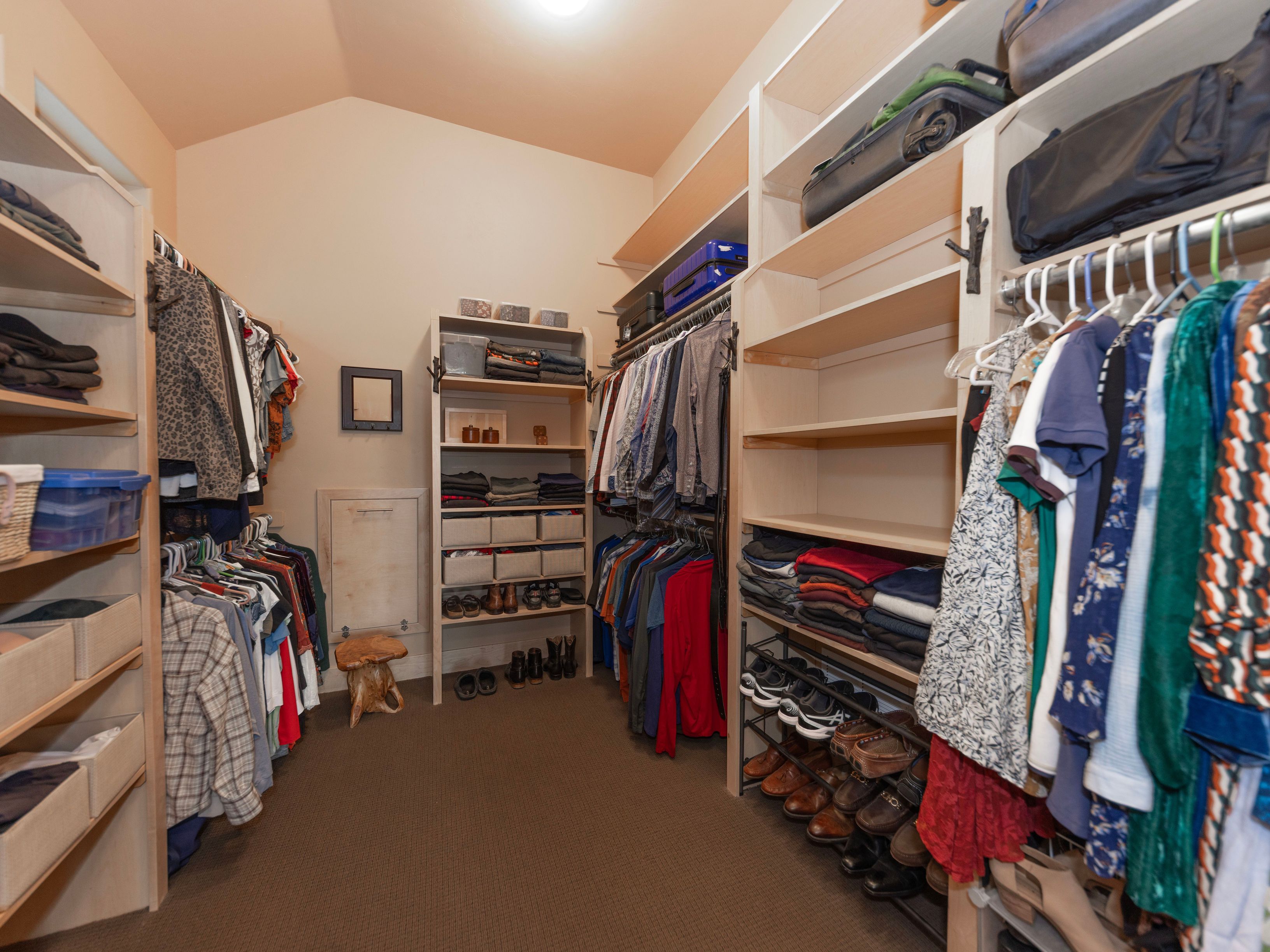 This spacious walk-in closet features extensive custom shelving and hanging rods on both sides, providing ample storage for clothing and accessories. The room is carpeted and includes a central shelving unit with a small wooden stool, creating a functional and organized dressing area. The neutral wall color and warm lighting contribute to a clean, well-maintained aesthetic suitable for a primary suite.
