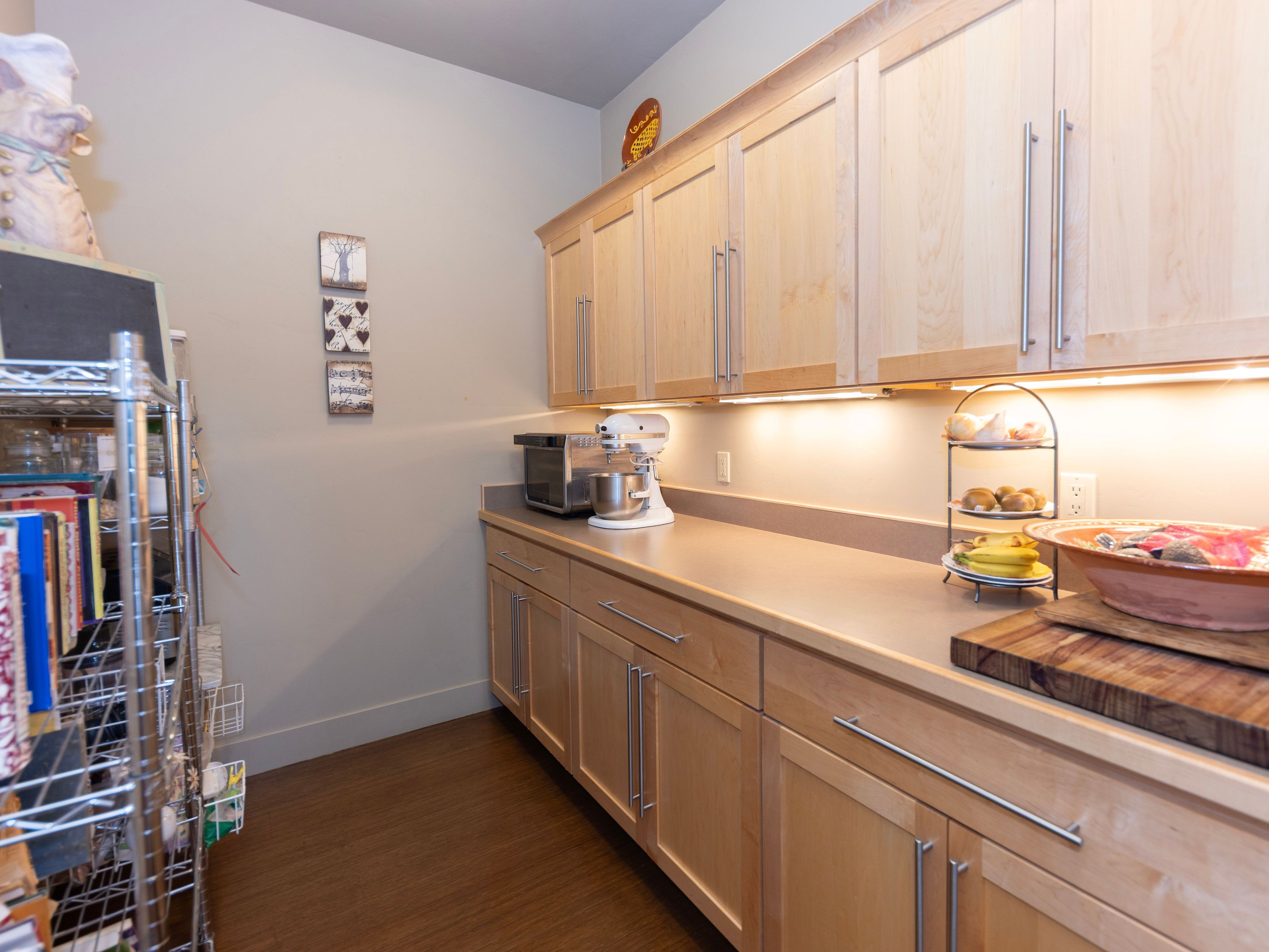 This kitchen area features light wood cabinetry with sleek silver hardware and a neutral countertop, providing a clean and functional workspace. A stand mixer sits on the counter alongside a tiered fruit stand, while a wire shelving unit on the left offers additional storage for books and pantry items. The space is illuminated by under-cabinet lighting, creating a warm and inviting atmosphere.