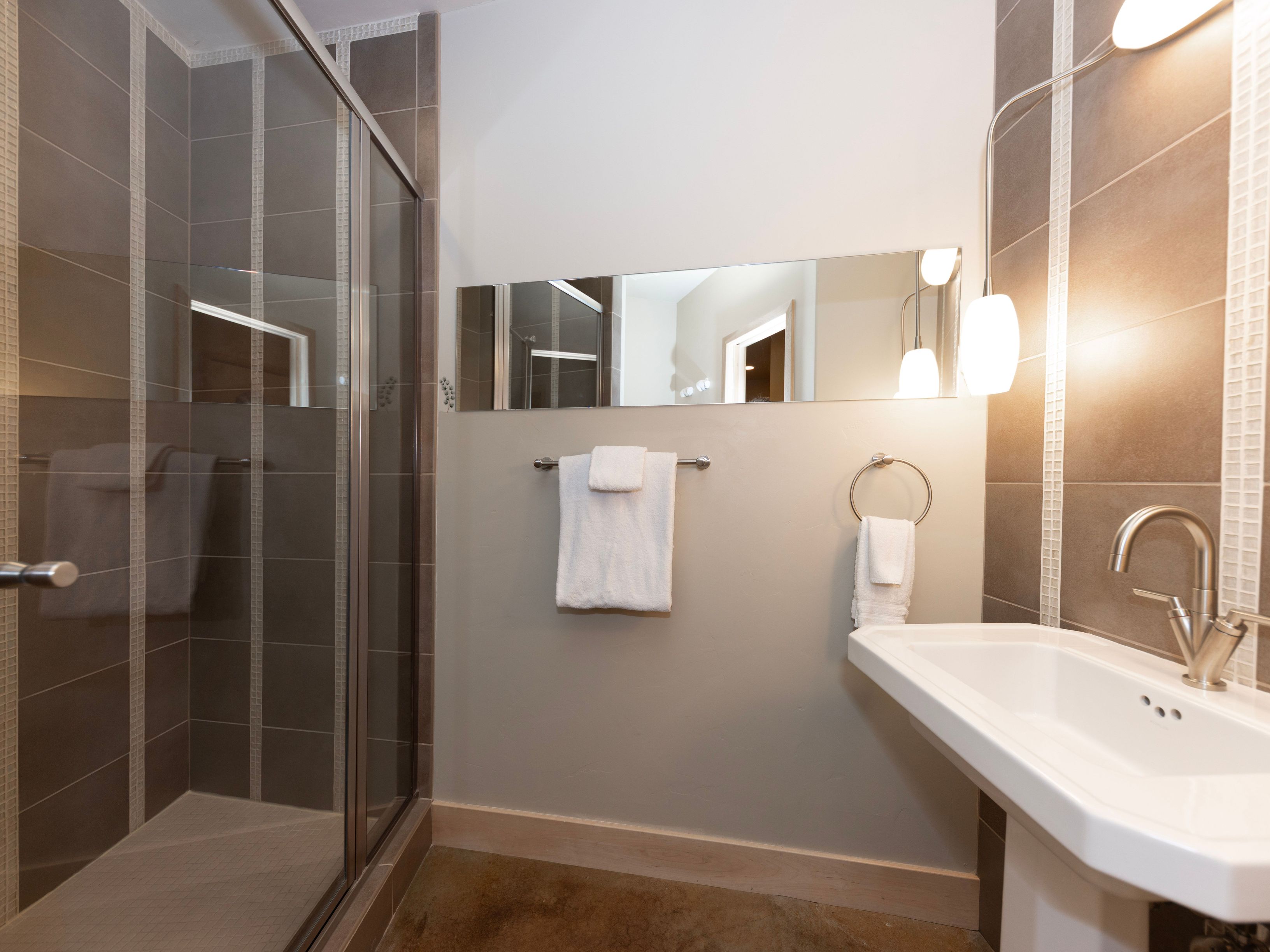 This modern bathroom features a glass-enclosed shower with neutral-toned tiled walls and a sleek pedestal sink. A long, horizontal mirror reflects the space, while a wall-mounted light fixture provides warm illumination above the vanity area. The room is accented with white towels hanging on a towel bar and a ring, creating a clean and functional aesthetic.
