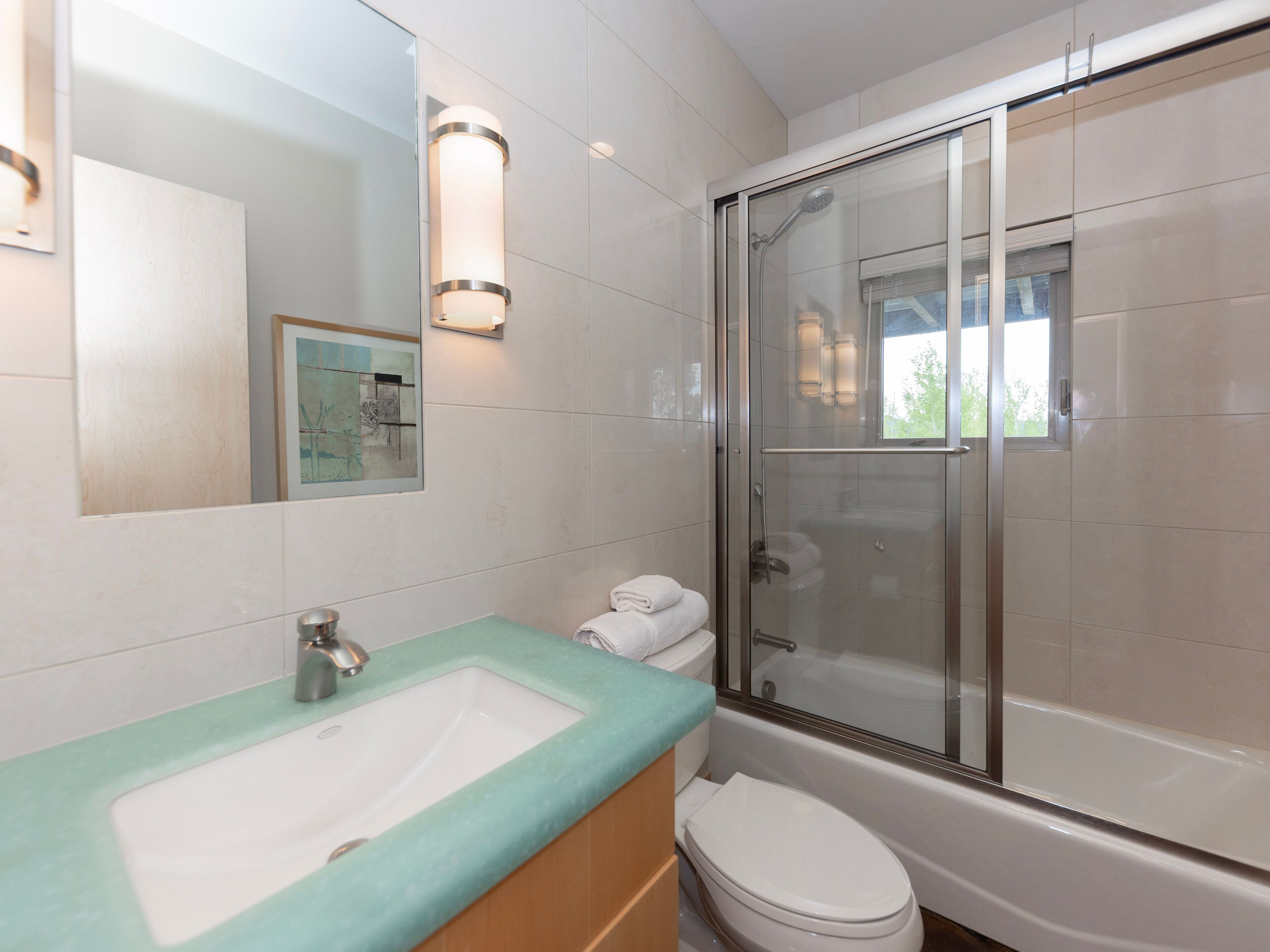 This compact bathroom features a vanity with a light-colored, translucent-style countertop and a simple chrome faucet. A glass-enclosed shower-tub combination is positioned next to a white toilet, with light-toned wall tiles throughout. The space is illuminated by a wall-mounted light fixture above the mirror, creating a clean and functional aesthetic.
