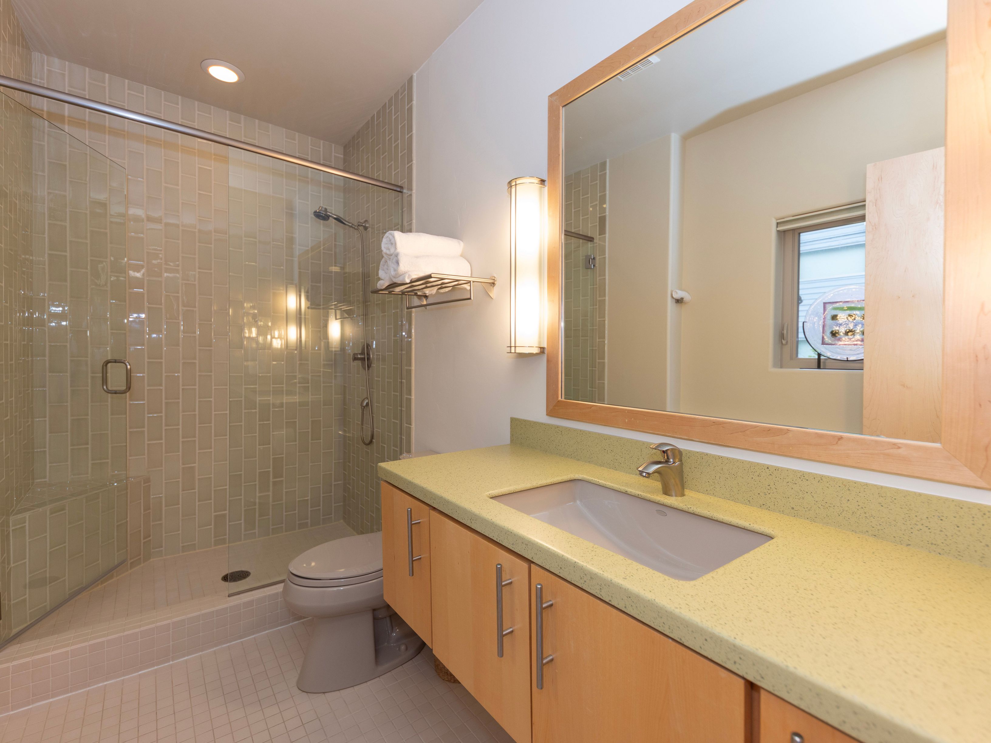 This clean and functional bathroom features a light wood vanity topped with a speckled stone counter and an integrated sink. A glass-enclosed shower with vertical subway-style tile walls sits adjacent to a neutral-toned toilet, while a large mirror with a wood frame reflects the space. The room is illuminated by a tall, vertical wall sconce and recessed lighting, creating a bright and well-maintained atmosphere.