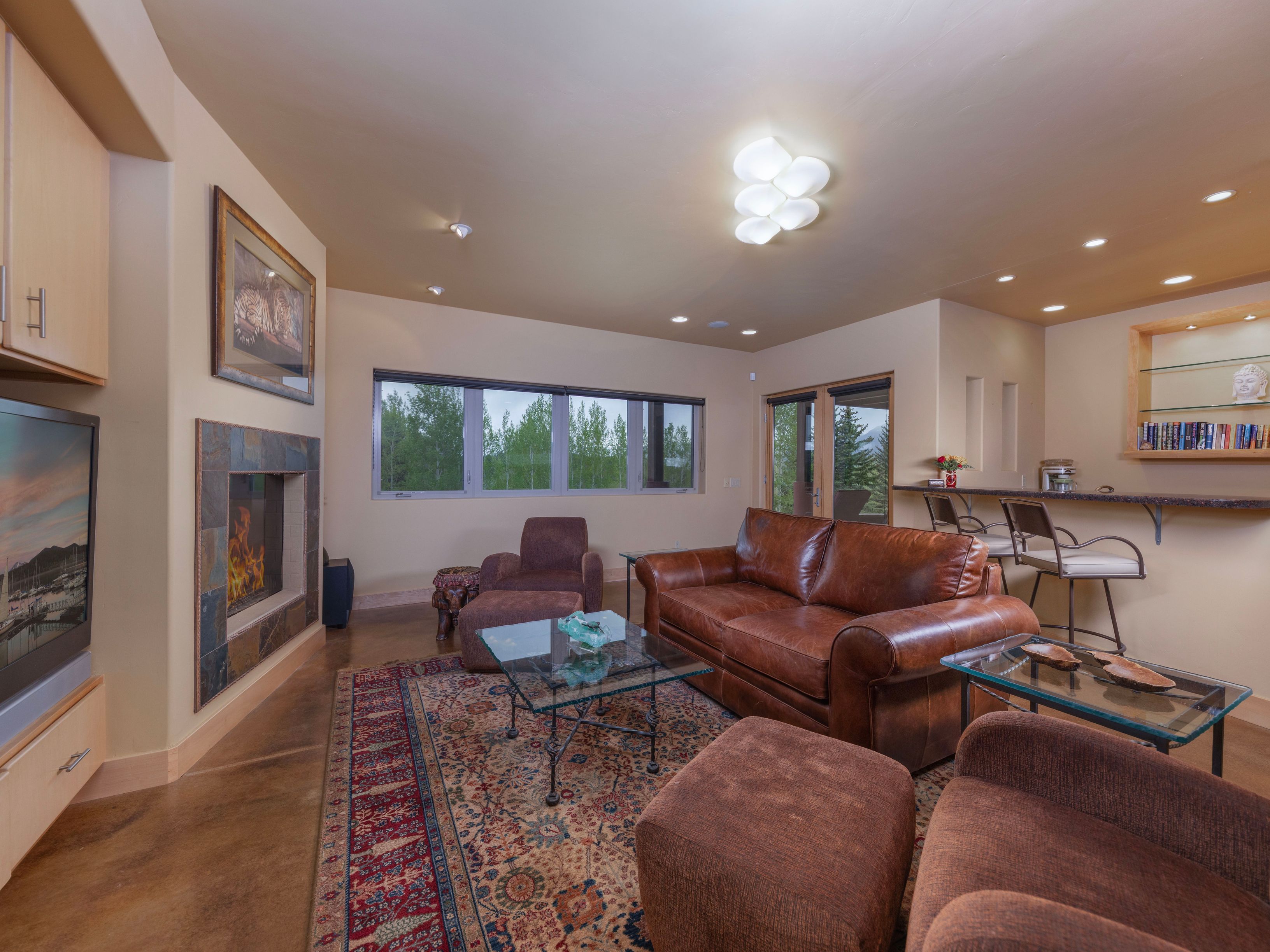This inviting living room features a warm, rustic aesthetic with a large leather sofa, matching armchairs, and a patterned area rug centered on a polished concrete floor. A stone-clad fireplace serves as a focal point, while a built-in bar area with stools and glass shelving provides a functional space for entertaining. Large windows offer a view of the surrounding trees, and the room is illuminated by a unique, multi-globe ceiling fixture.