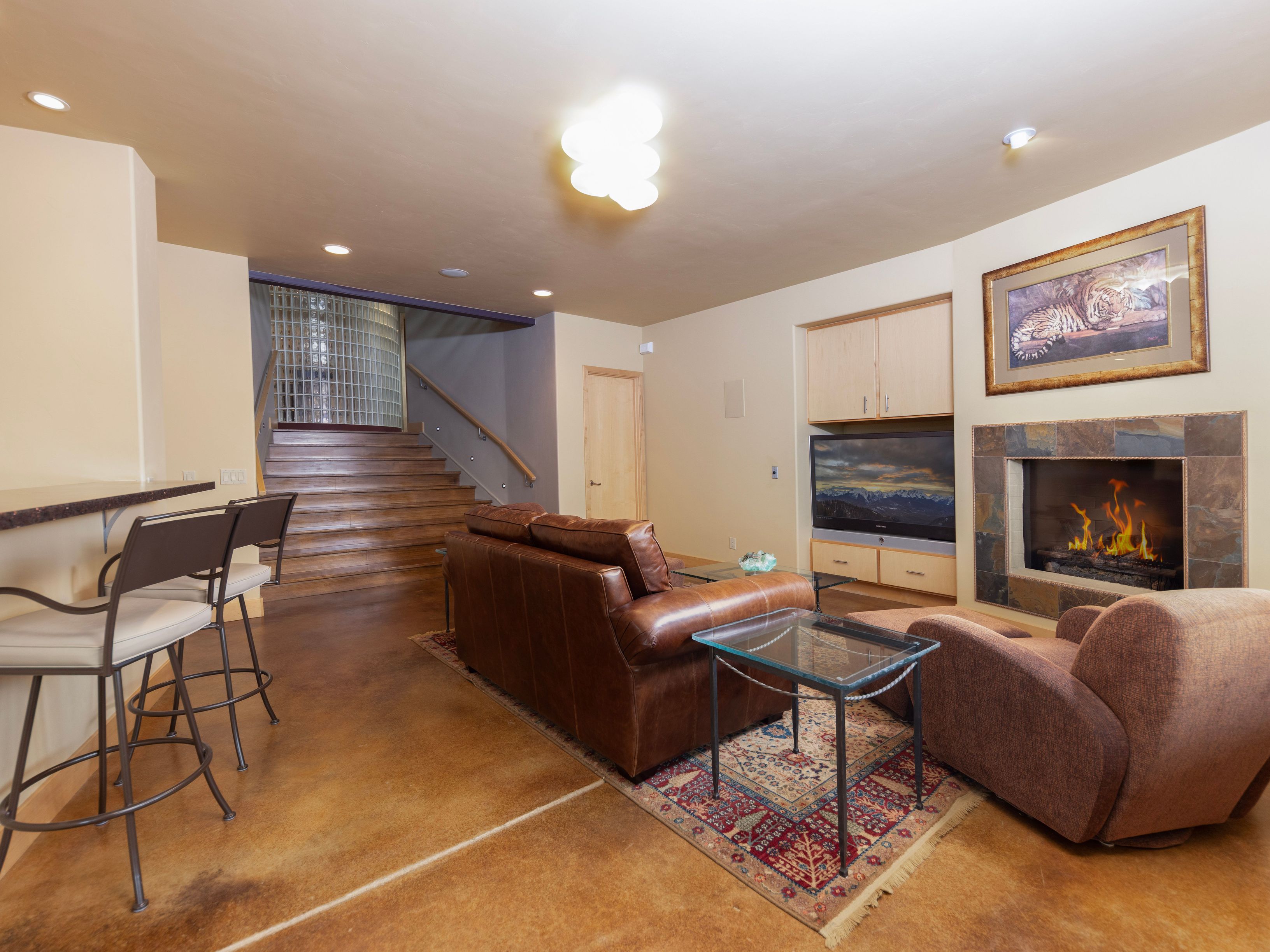 This spacious basement living area features a warm, inviting atmosphere with a brown leather sofa, a plush armchair, and a glass-topped side table resting on a patterned area rug. A stone-clad fireplace serves as a focal point, complemented by a built-in media cabinet and a bar area with high stools. The room is anchored by polished concrete floors and leads to a staircase with a glass-block wall, creating a comfortable and functional entertainment space.