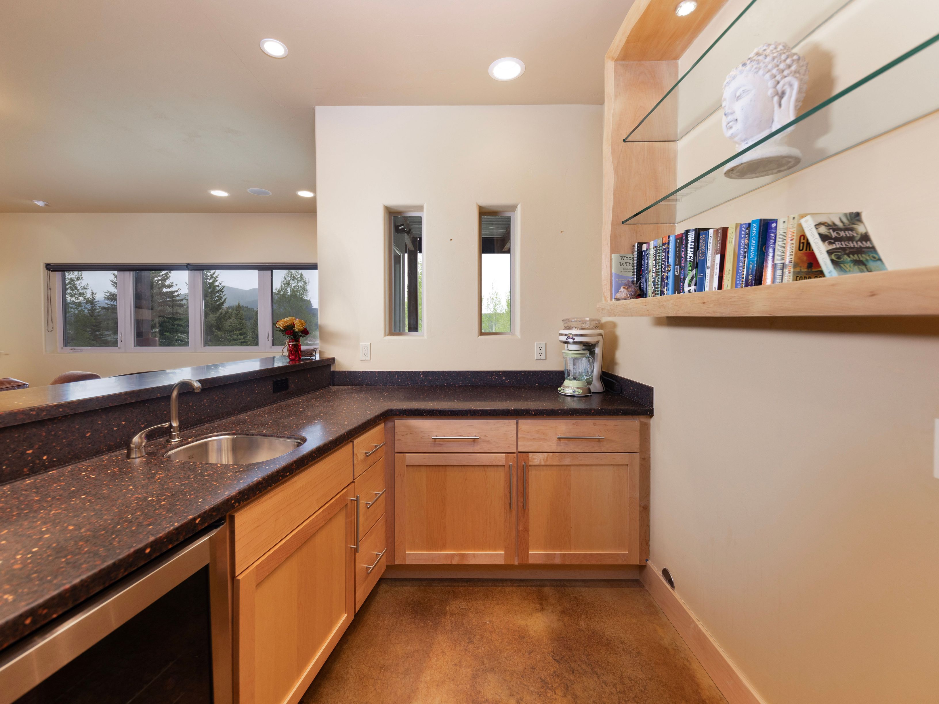 This kitchen features light wood cabinetry paired with dark, speckled countertops and a stainless steel sink. A built-in shelving unit with glass tiers displays books and a decorative bust, while two narrow windows provide natural light. The space is finished with a smooth, warm-toned floor, creating a functional and organized culinary area.