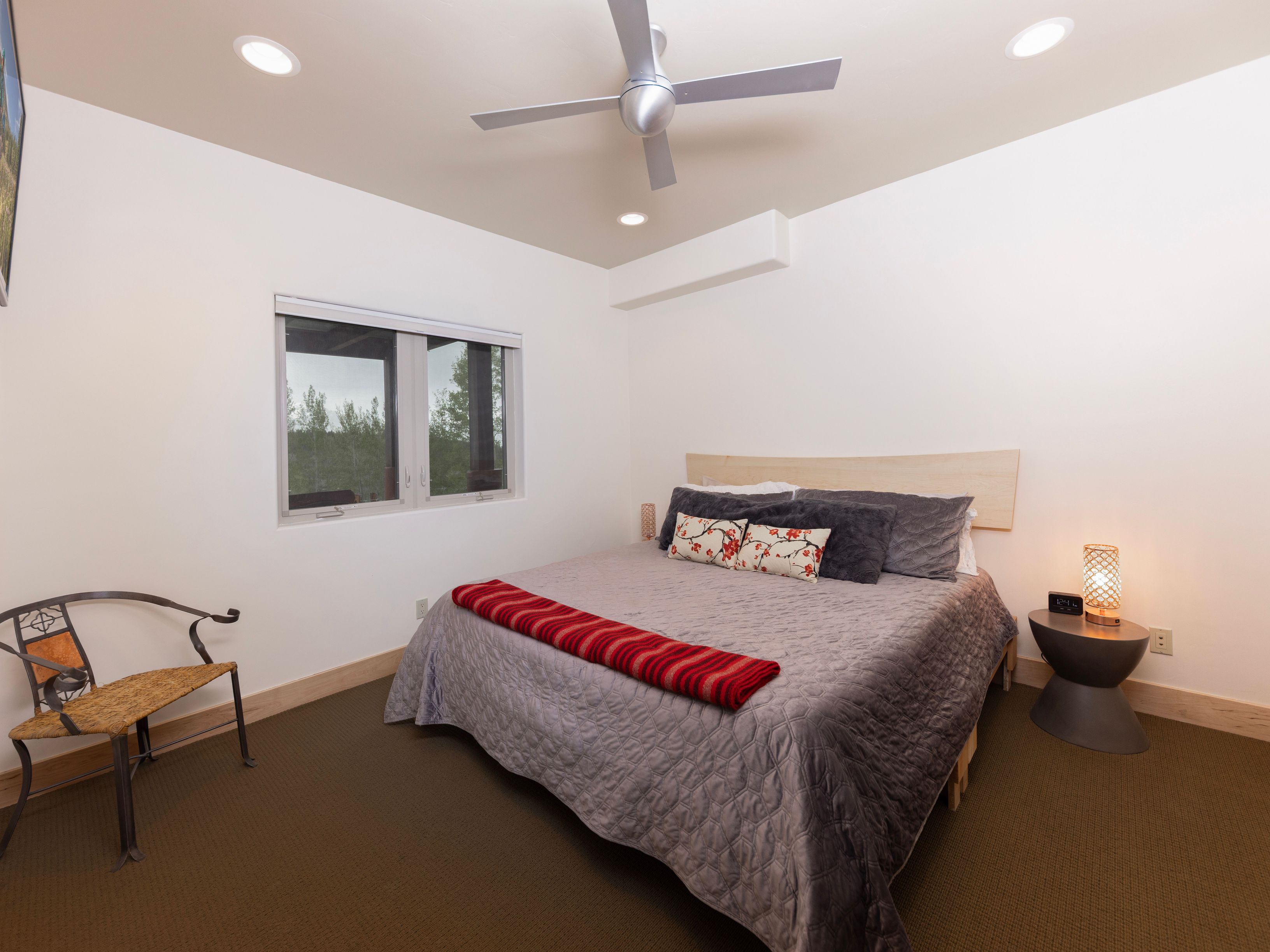 This bedroom features a neutral color palette with a queen-sized bed dressed in a quilted gray coverlet and a vibrant red-striped throw blanket. The room is accented by a unique metal-framed chair with a woven seat and a modern bedside table topped with a textured lamp. A window provides natural light and a view of the surrounding trees, while a ceiling fan and recessed lighting complete the space.