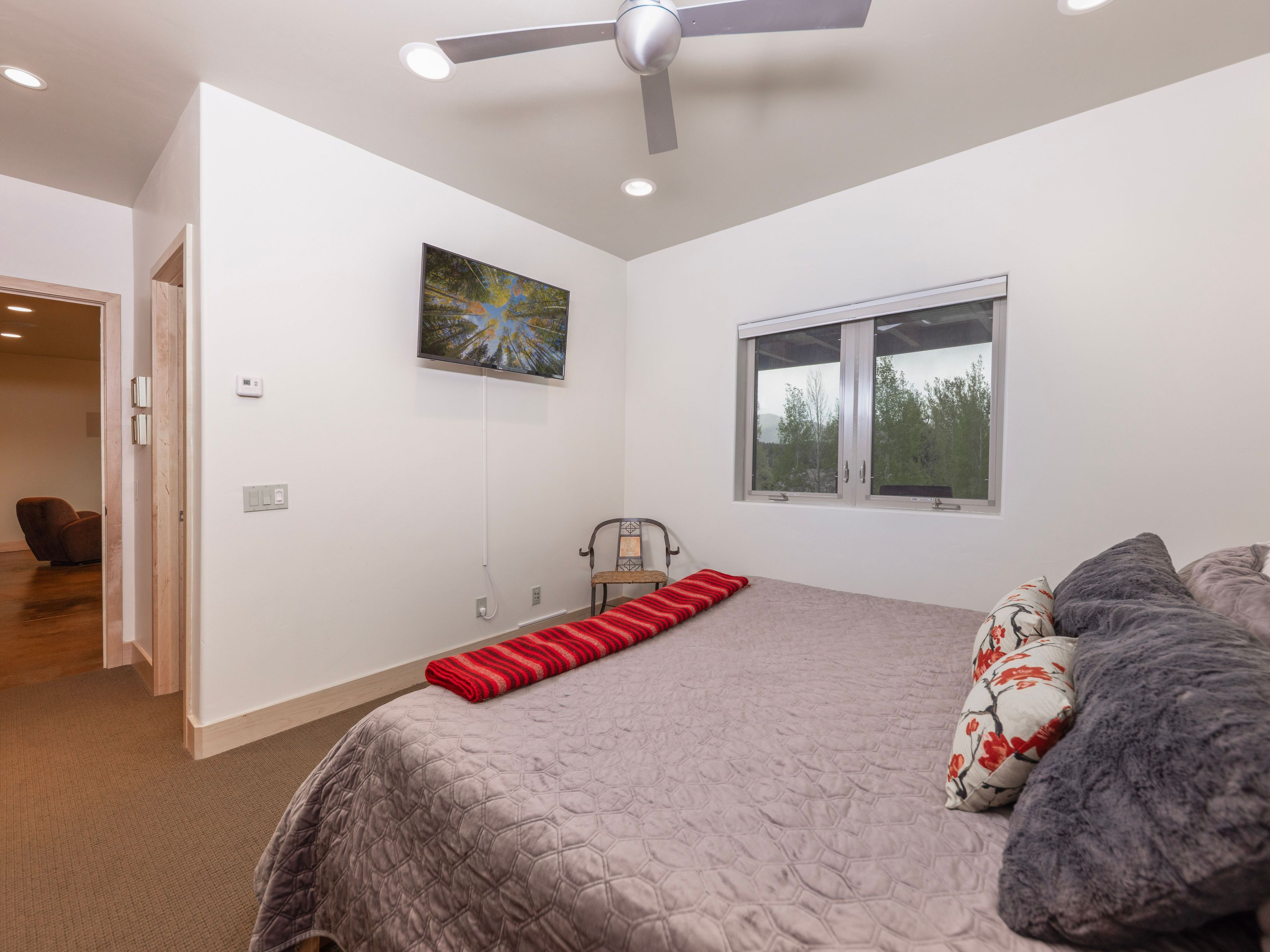 This bedroom features a neutral color palette with a textured gray bedspread and decorative floral pillows. A wall-mounted television is positioned above a small, vintage-style metal chair, while a red patterned throw adds a pop of color to the foot of the bed. The room is illuminated by recessed lighting and a modern ceiling fan, with a window providing a view of the surrounding trees.