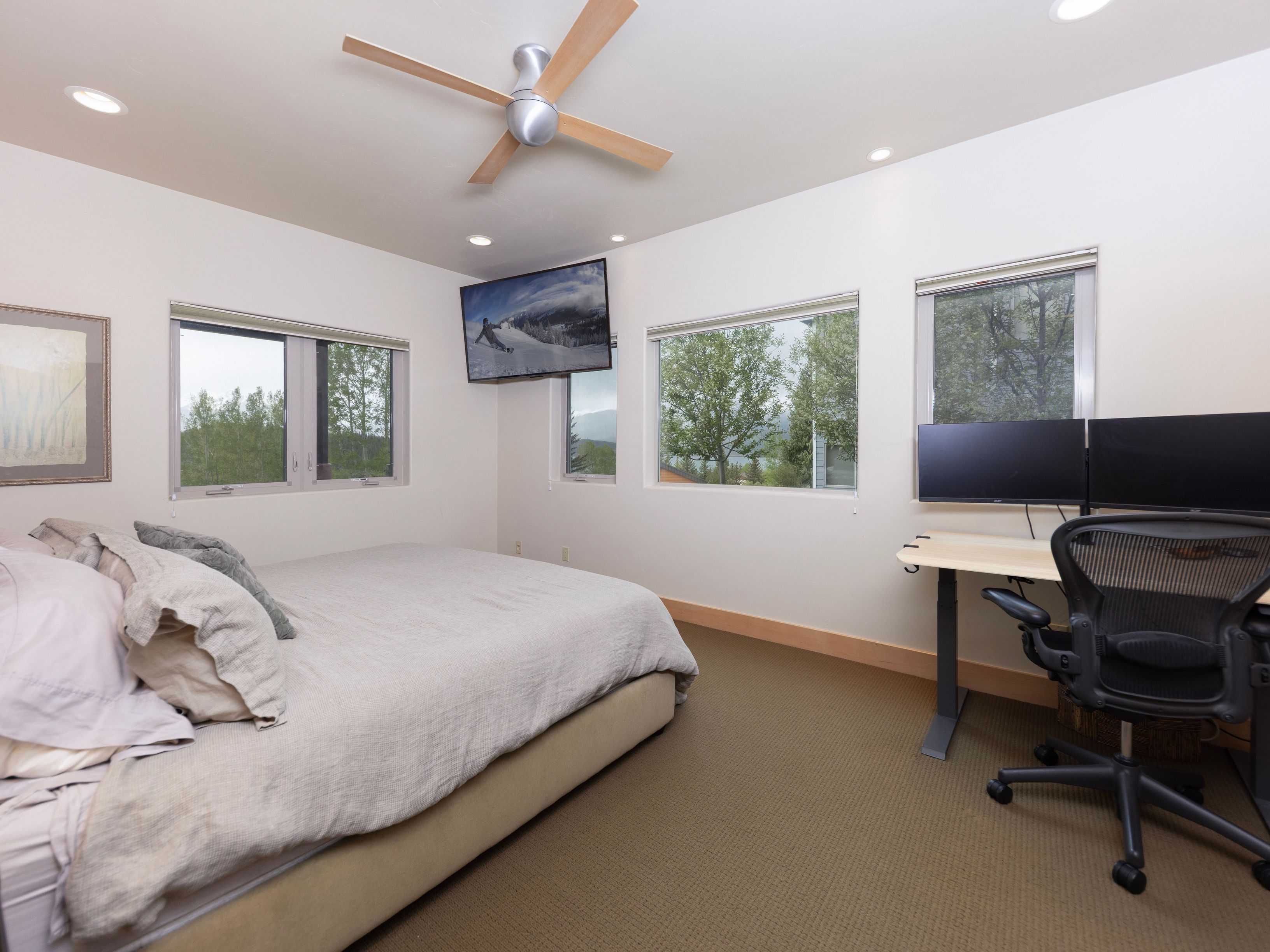 This multi-functional bedroom features a neutral color palette with a comfortable bed and a dedicated workspace equipped with a standing desk and dual monitors. Large windows provide ample natural light and views of the surrounding trees, while a ceiling fan and wall-mounted television add convenience to the space. The room is carpeted and maintains a clean, practical aesthetic suitable for both rest and productivity.