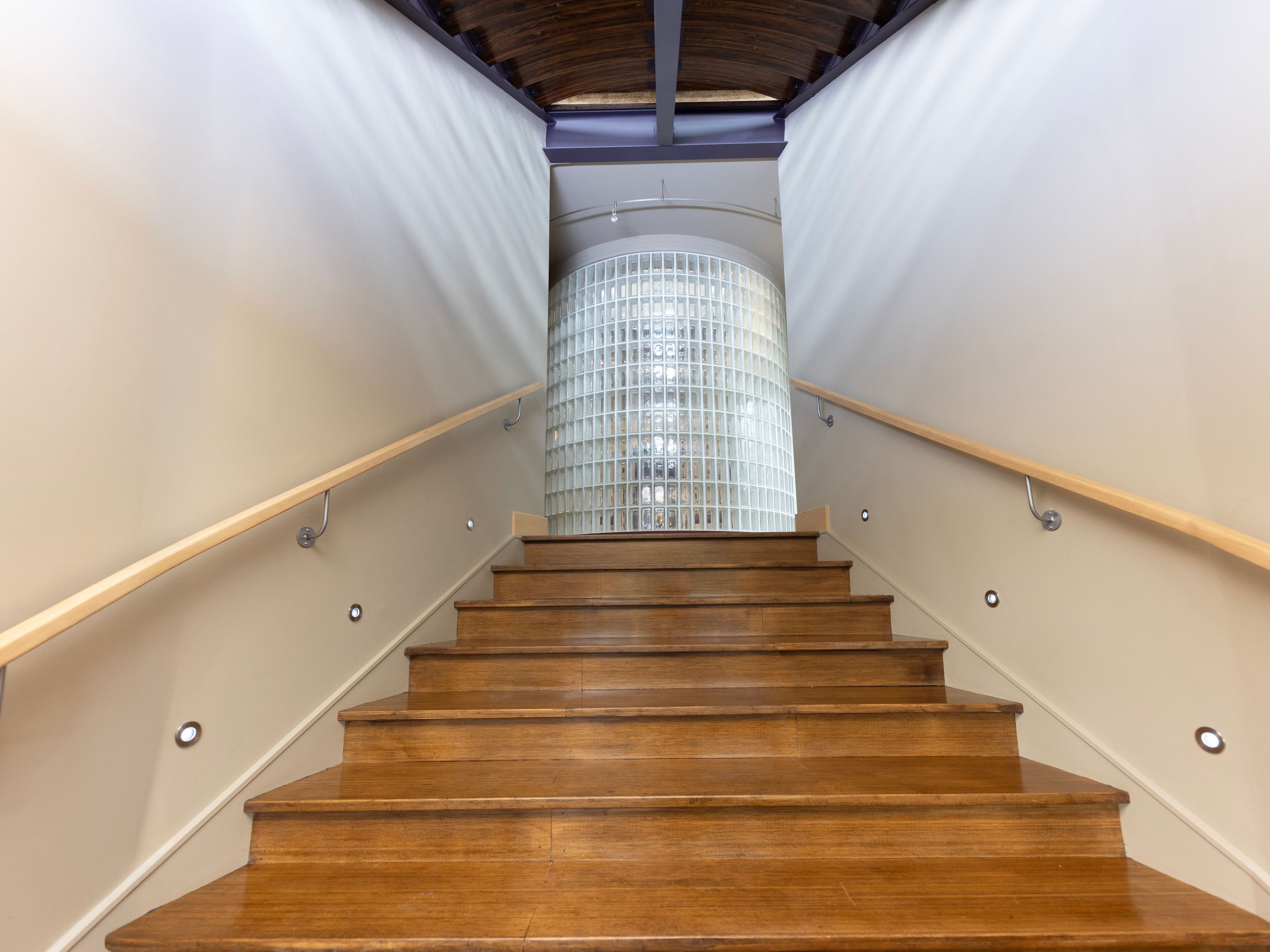 This perspective captures a set of wooden stairs ascending toward a striking, curved glass-block wall that serves as a focal point. The staircase is flanked by light-colored walls, simple wooden handrails, and recessed accent lighting embedded along the path. The ceiling features a dark, arched wooden structure, creating a warm and architectural transition between levels.