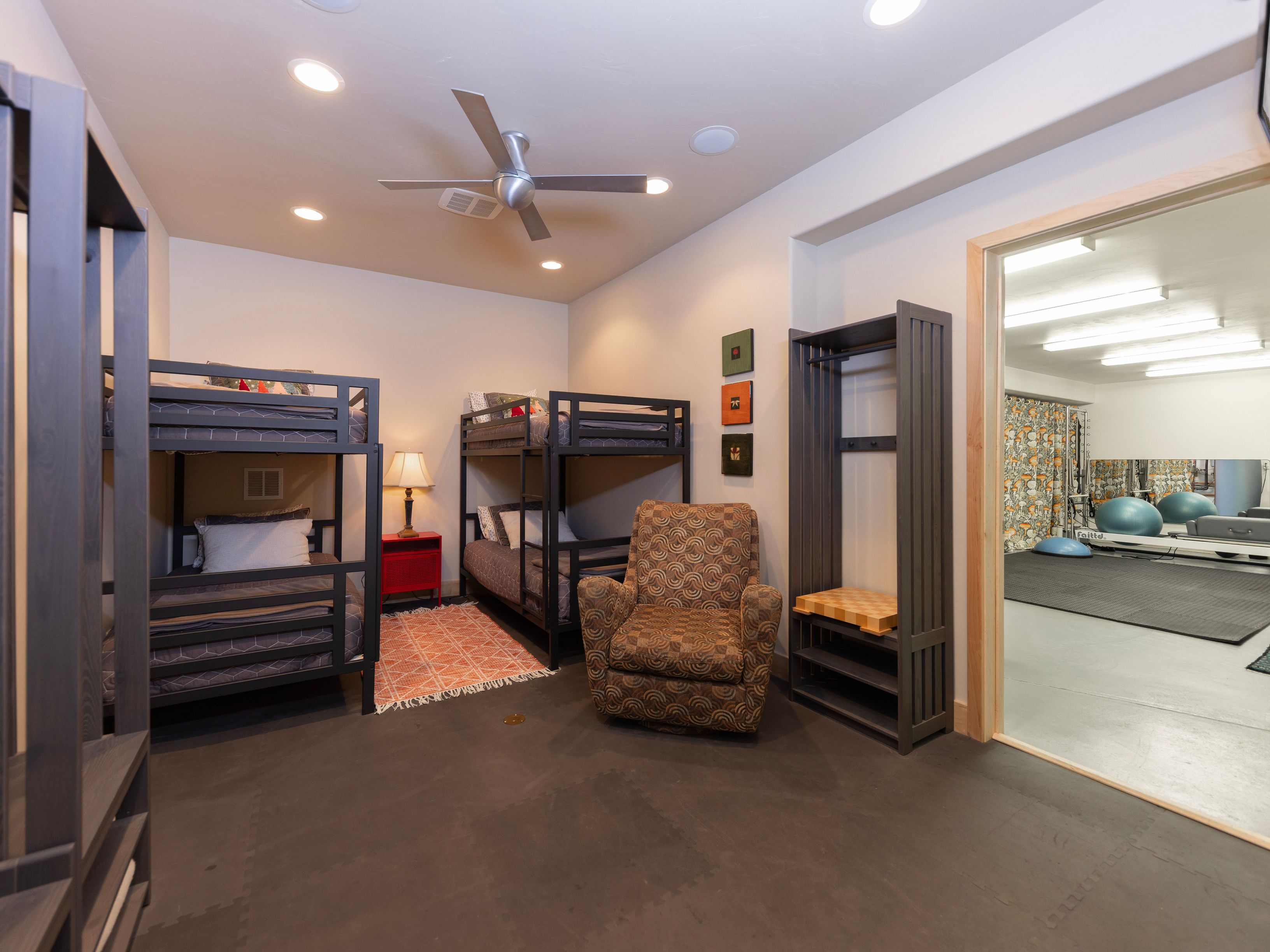 This versatile bedroom features two dark-framed bunk beds, providing ample sleeping space, and a patterned armchair for comfortable seating. A small red nightstand with a lamp sits between the beds, while an open-frame storage unit stands near the doorway leading to an adjacent home gym. The room has a neutral color palette with dark flooring and recessed lighting, creating a functional and multi-purpose living area.