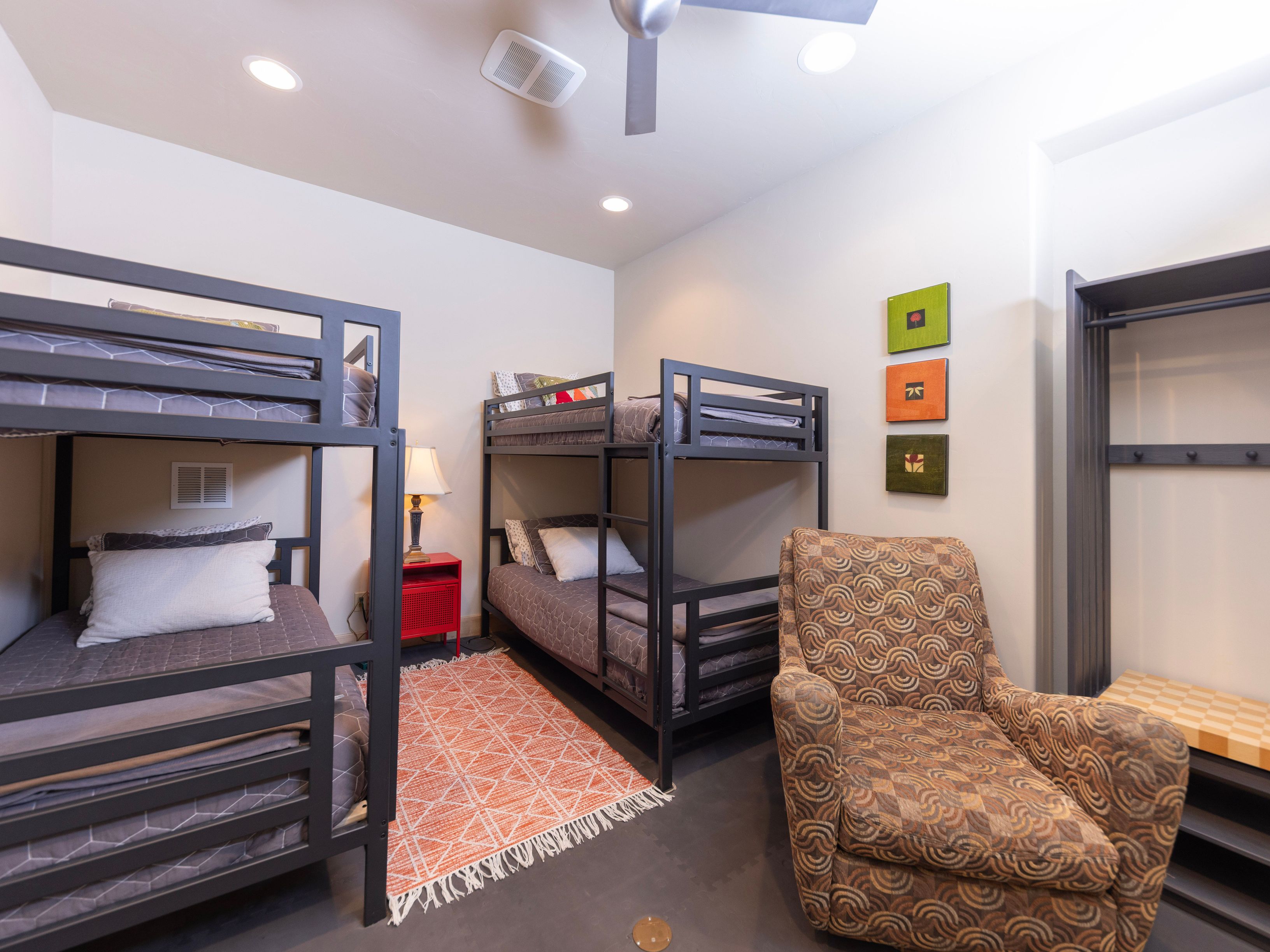 This functional bedroom features two dark metal bunk beds arranged to maximize sleeping capacity, complemented by a patterned area rug and a patterned armchair. A small red nightstand with a lamp sits between the beds, while a wall-mounted closet unit provides storage space. The room is decorated with three small square art pieces, creating a cozy and efficient space suitable for guests or children.