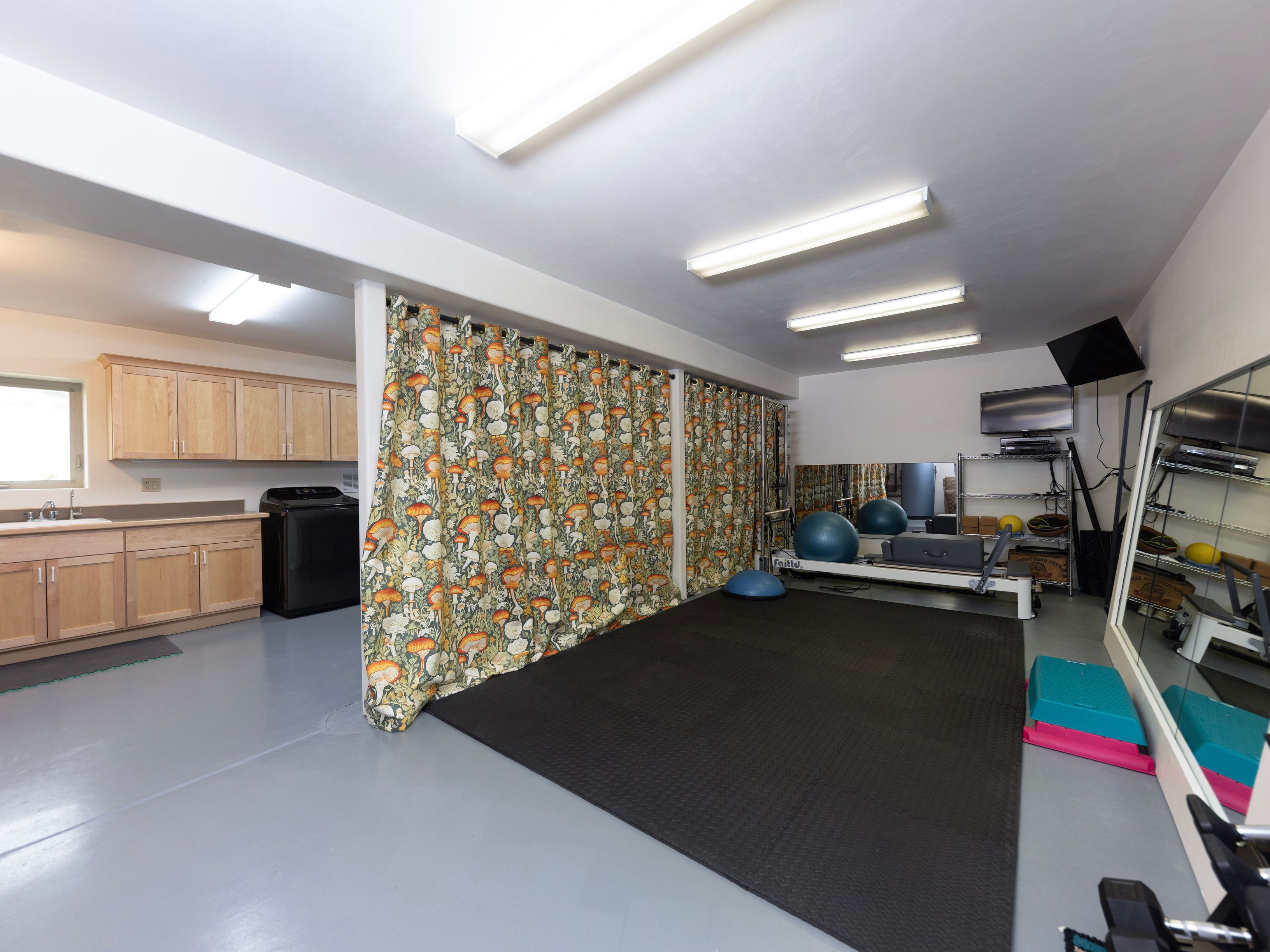 This versatile space functions as a home gym, featuring a large black rubber mat for exercise equipment, including a Pilates reformer and balance balls. A patterned curtain with a botanical mushroom motif acts as a room divider, separating the workout area from a laundry zone equipped with light wood cabinetry and a sink. The room is illuminated by multiple linear ceiling lights and includes a large wall mirror that enhances the sense of space.