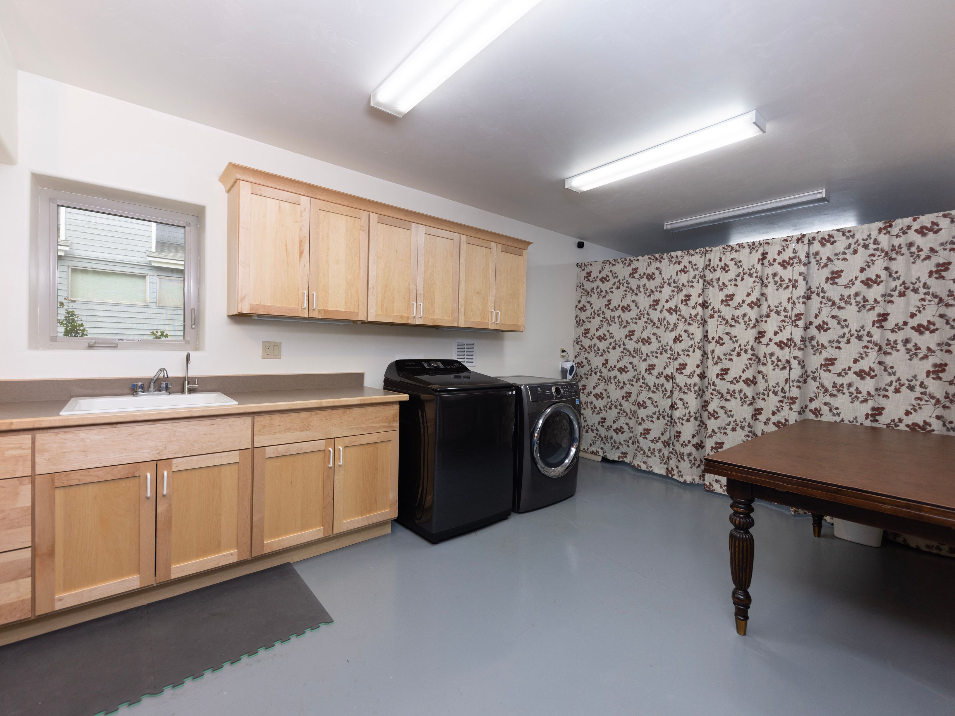 This functional laundry room features light wood cabinetry providing ample storage above a sink and countertop area. A black washer and dryer set are positioned centrally, while a patterned curtain covers a large section of the wall, adding a decorative touch. The space is illuminated by two long, rectangular ceiling-mounted light fixtures, and a wooden table sits in the foreground, suggesting a versatile workspace.