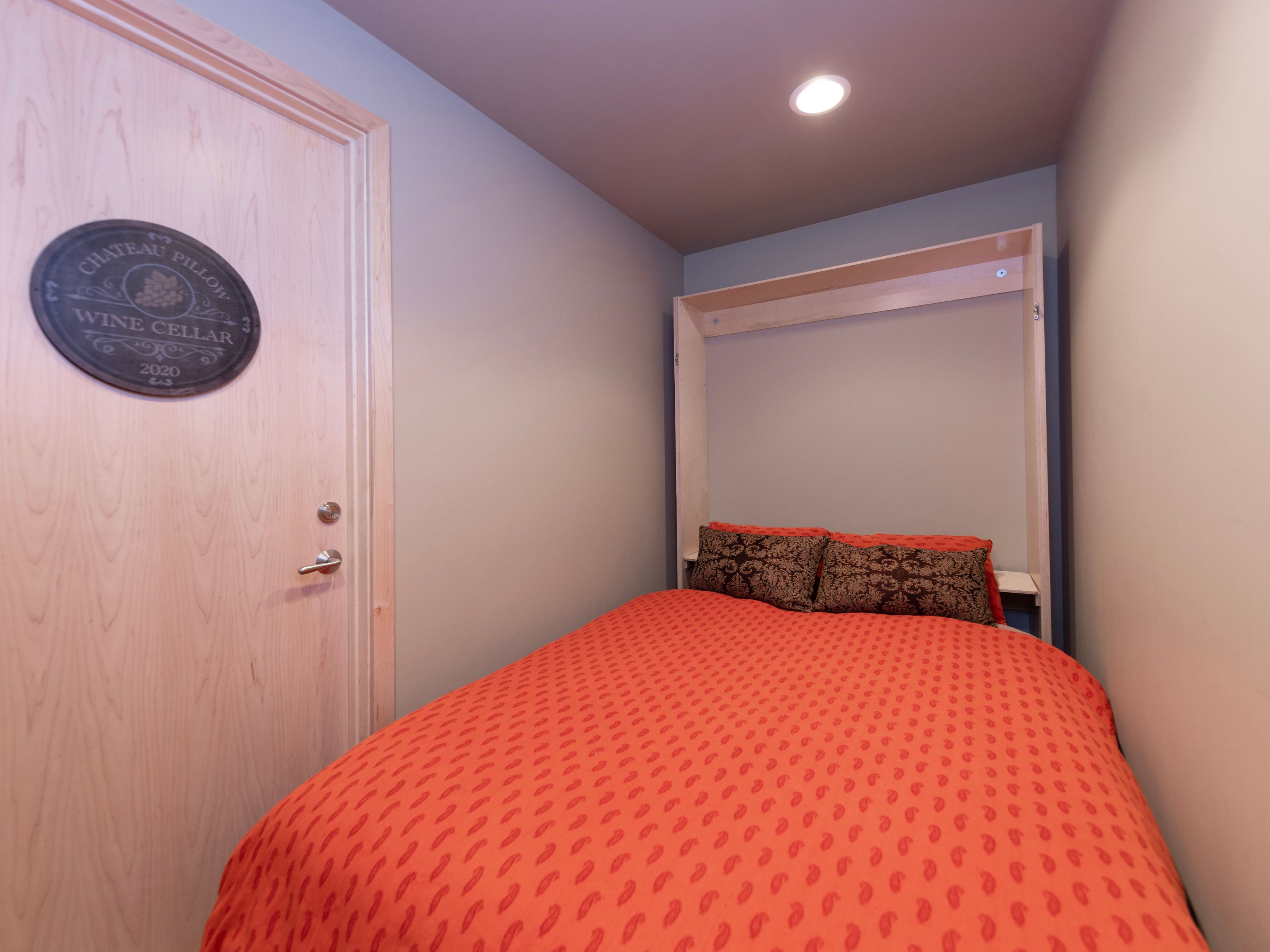 This cozy guest bedroom features a space-saving Murphy bed frame in a light wood finish, topped with a vibrant orange patterned duvet and two dark, textured accent pillows. To the left, a light wood door is adorned with a decorative 'Chateau Pillow Wine Cellar' sign, adding a touch of rustic charm to the room. The perspective is a straight-on shot that highlights the efficient use of space in this compact, neutrally painted bedroom.