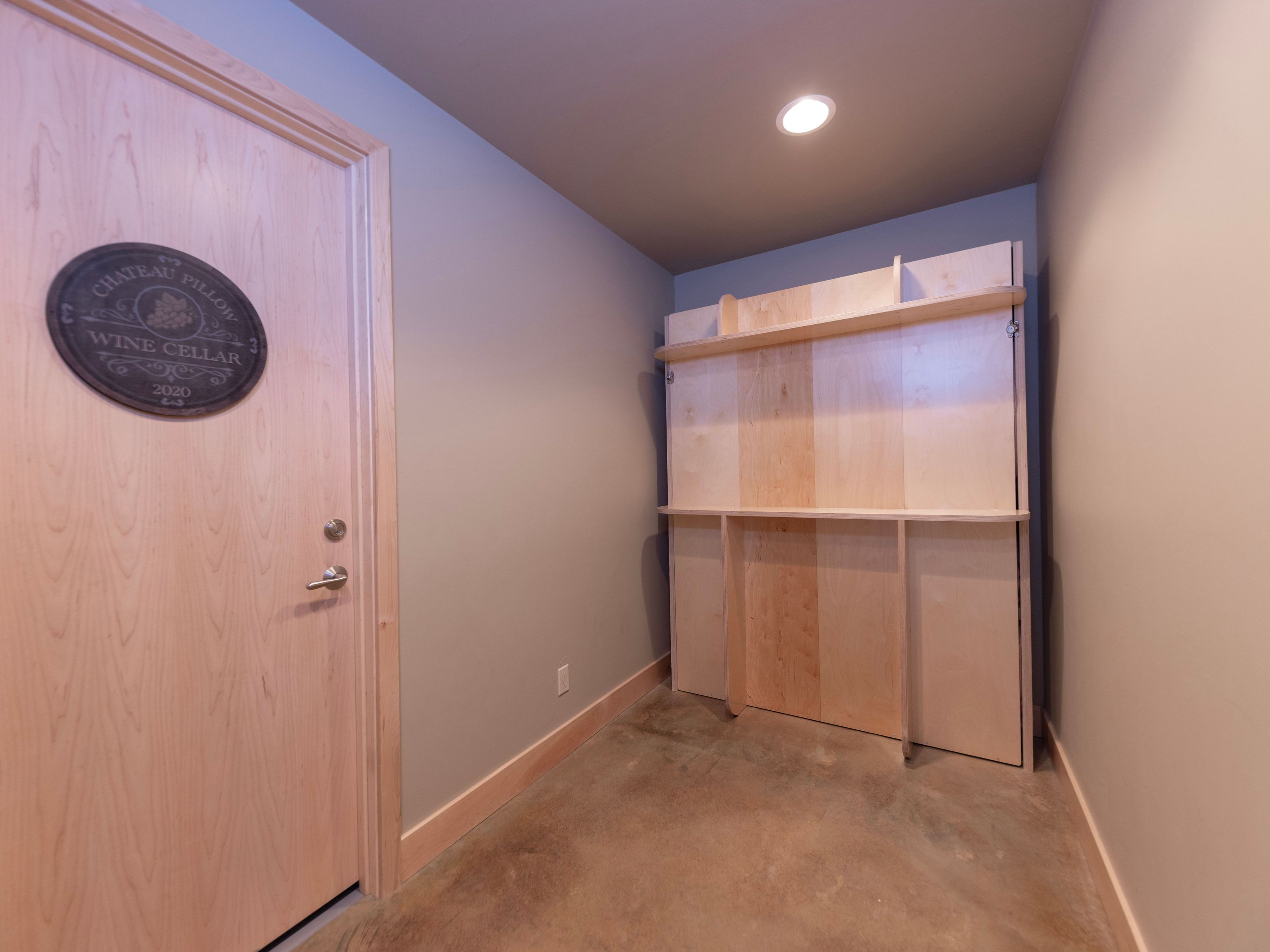 This interior space features a light wood door with a decorative 'Chateau Pillow Wine Cellar 2020' plaque, suggesting a dedicated storage area. The room includes a custom-built light wood shelving unit against a neutral-toned wall, set above a simple concrete floor. The perspective is a straight-on view, highlighting the minimalist and functional design of the storage area.