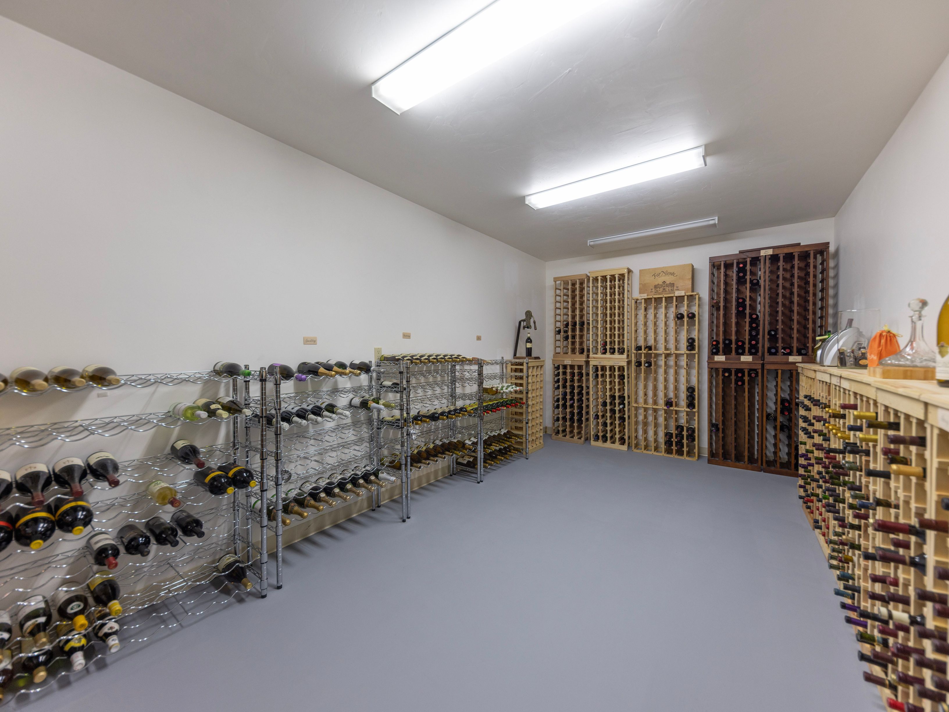 This dedicated wine cellar features extensive storage solutions, including metal wire racking along the left wall and wooden grid shelving units filling the back and right walls. The room has a clean, minimalist aesthetic with light-colored walls and a smooth, neutral-toned floor. The perspective is a wide-angle shot from the entrance, showcasing the organized collection and the functional layout of the space.