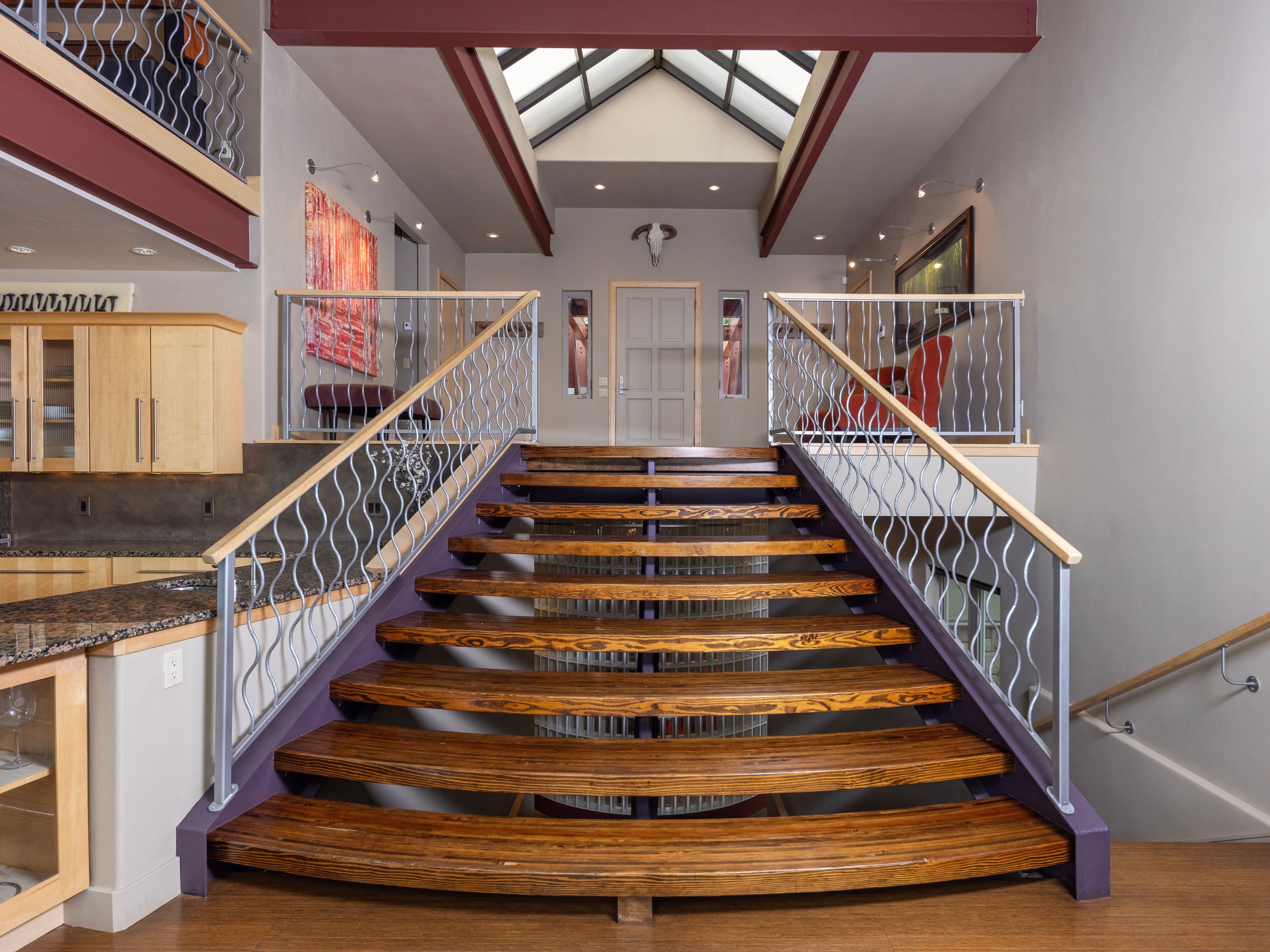This grand entryway features a prominent central staircase with rich, dark wood treads and a distinctive wavy metal railing. The space is illuminated by a large skylight above, casting natural light down onto the landing and the front door, which is adorned with a decorative animal skull. To the left, a partial view of a kitchen with light wood cabinetry and granite countertops adds functional context to this open-concept architectural design.