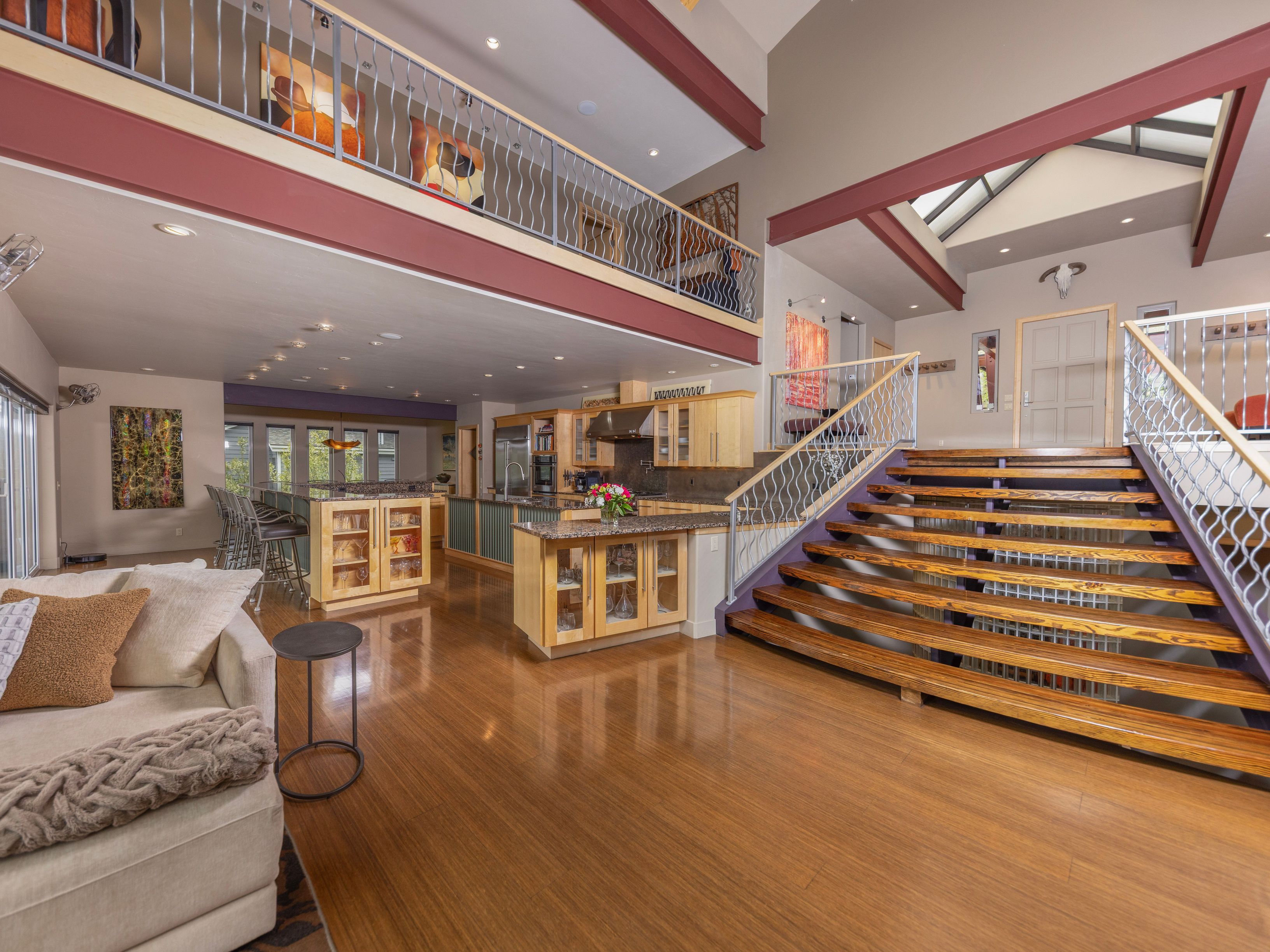 This expansive, open-concept living area features a high-ceiling design with a mezzanine level overlooking the main floor. The space is anchored by a prominent wooden staircase with metal railings, leading to the upper level, while the kitchen area is visible in the background with light wood cabinetry and a central island. The polished wood flooring reflects the ambient light, creating a warm and inviting atmosphere throughout this contemporary home.