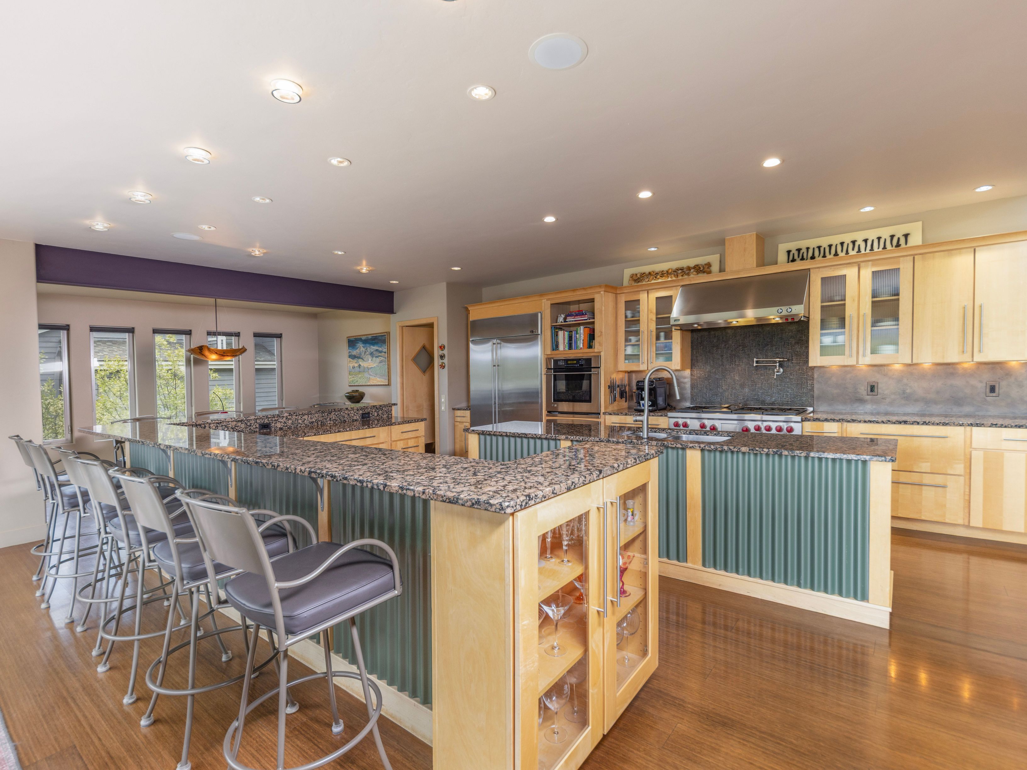 This spacious, open-concept kitchen features a large L-shaped island with a granite countertop and a unique corrugated metal base, providing ample seating for four. The cabinetry is crafted from light wood, complemented by stainless steel appliances and a professional-grade range hood. The perspective captures the expansive layout, highlighting the seamless flow between the cooking area and the breakfast bar, all set against a backdrop of warm wood flooring and recessed lighting.