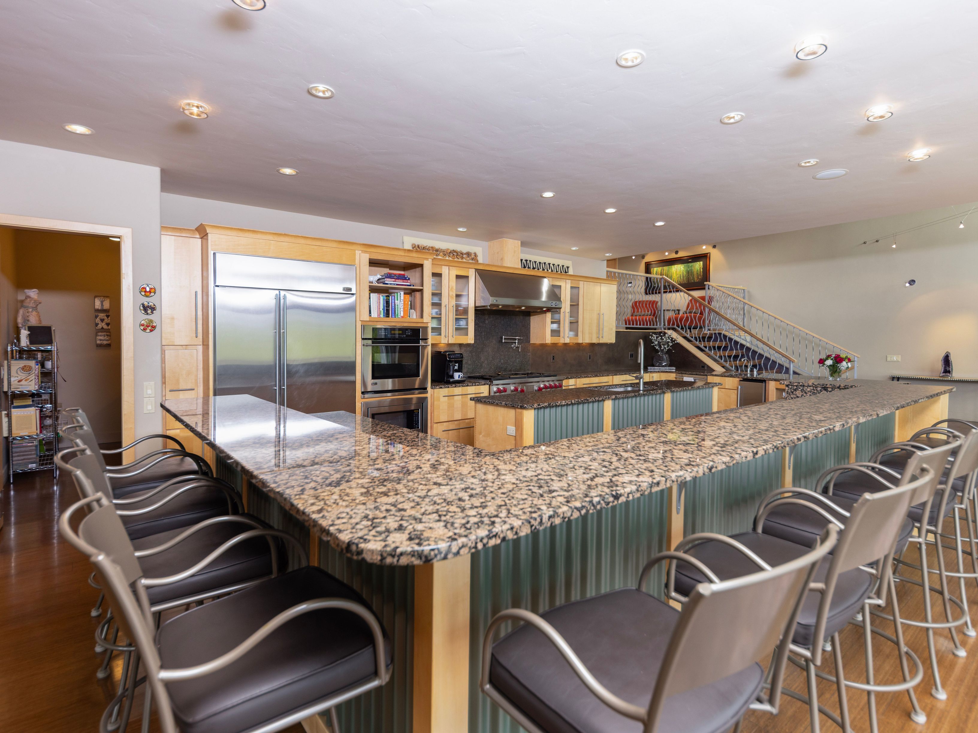 This spacious, open-concept kitchen features a large, L-shaped island with a speckled stone countertop and a corrugated metal base, providing ample seating with multiple bar stools. The cabinetry is finished in a light wood tone, complemented by stainless steel appliances, including a built-in refrigerator and wall oven. The perspective captures the expansive layout, leading the eye toward a staircase in the background, creating a bright and functional living space.