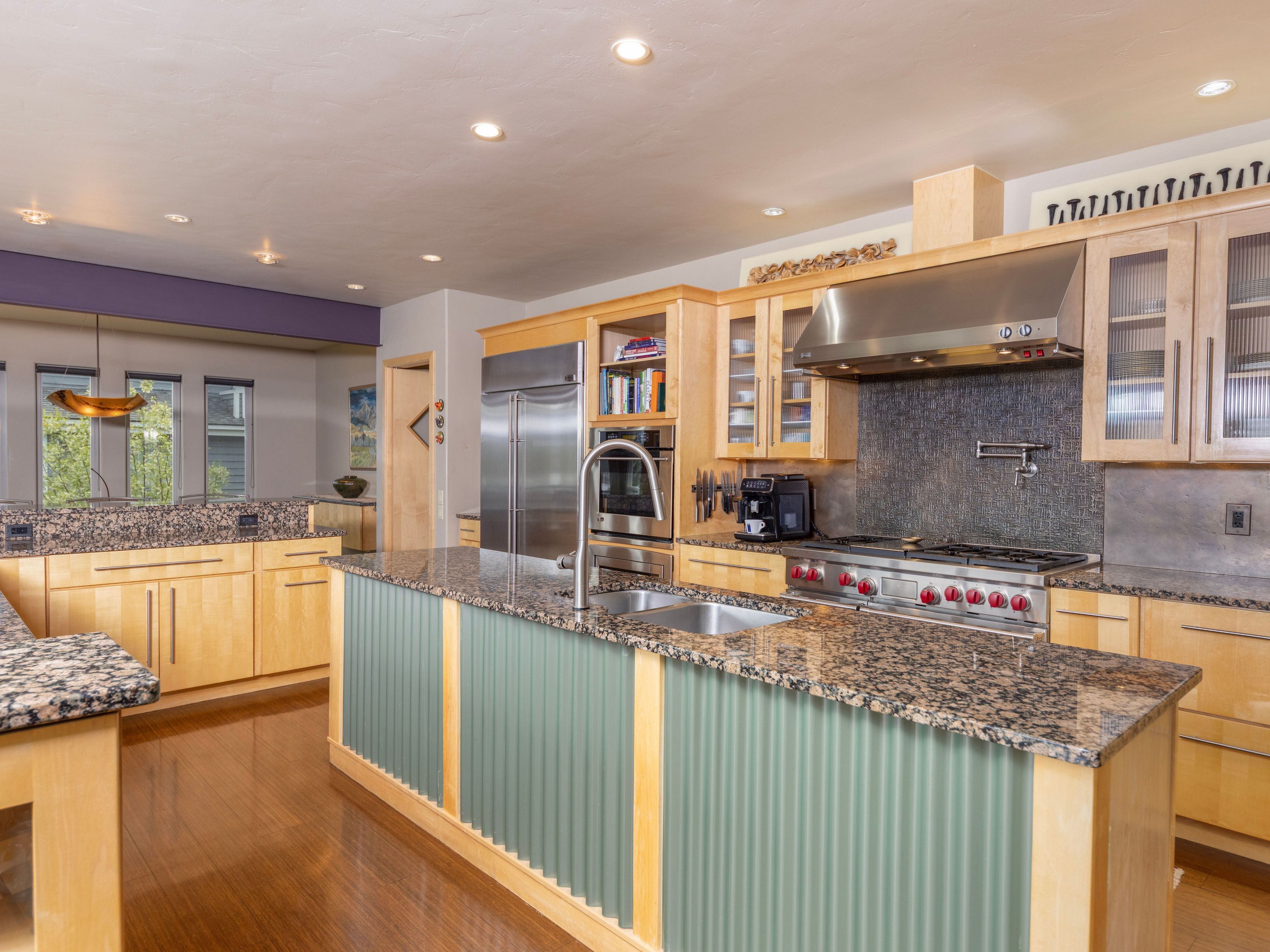 This spacious kitchen features light wood cabinetry, granite countertops, and a prominent central island with a unique corrugated metal front panel. The space is equipped with high-end stainless steel appliances, including a professional-style range and a large refrigerator, complemented by a dark tiled backsplash. The perspective captures the open-concept layout, highlighting the warm wood flooring and recessed lighting that creates an inviting, functional atmosphere.