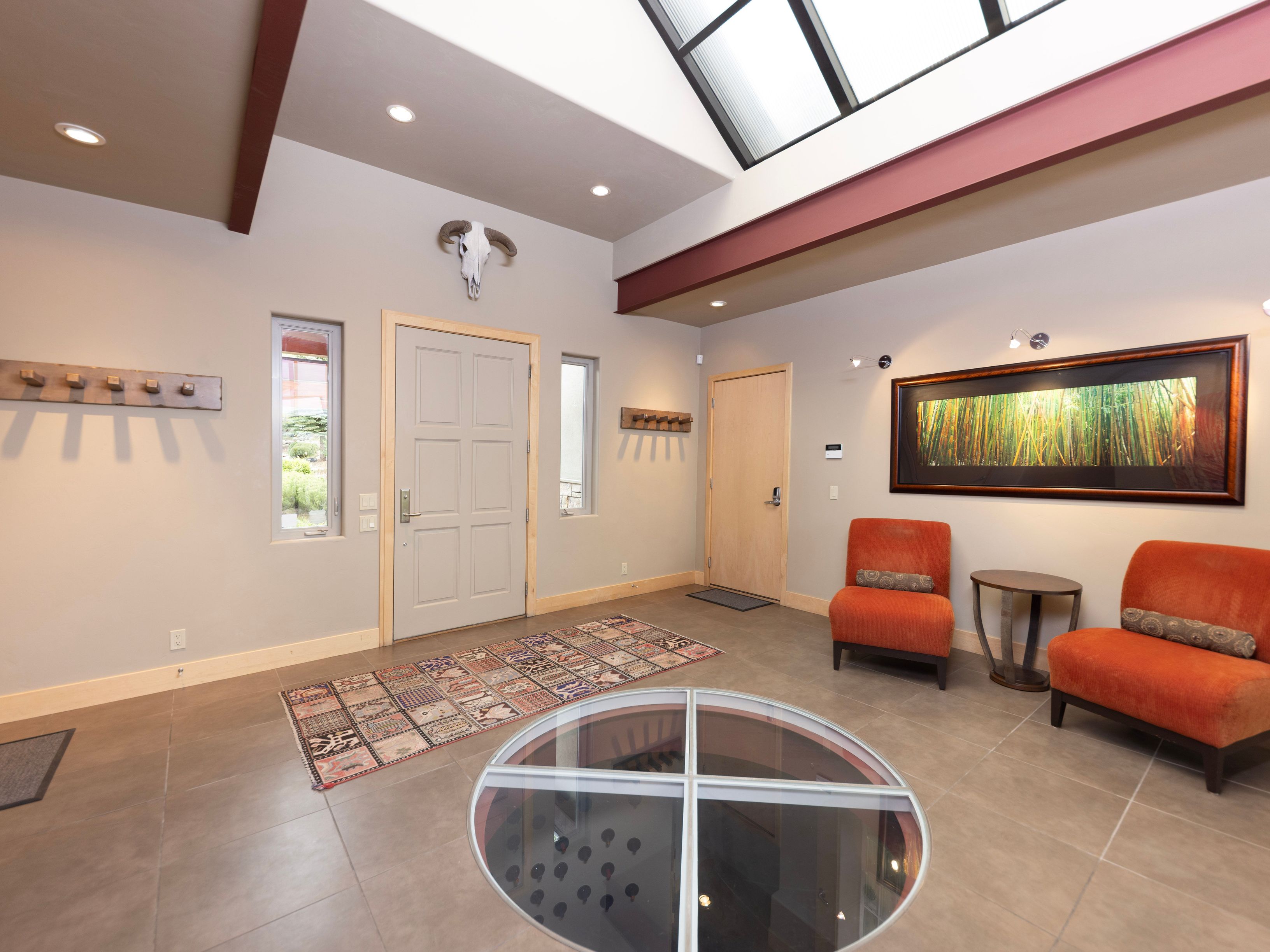 This spacious, modern entryway features a unique circular glass floor insert that provides a view into the level below. The room is accented by a skull wall mount, a patterned area rug, and two vibrant orange armchairs flanking a small side table. A large skylight overhead floods the neutral-toned space with natural light, highlighting the clean lines and contemporary architectural design.