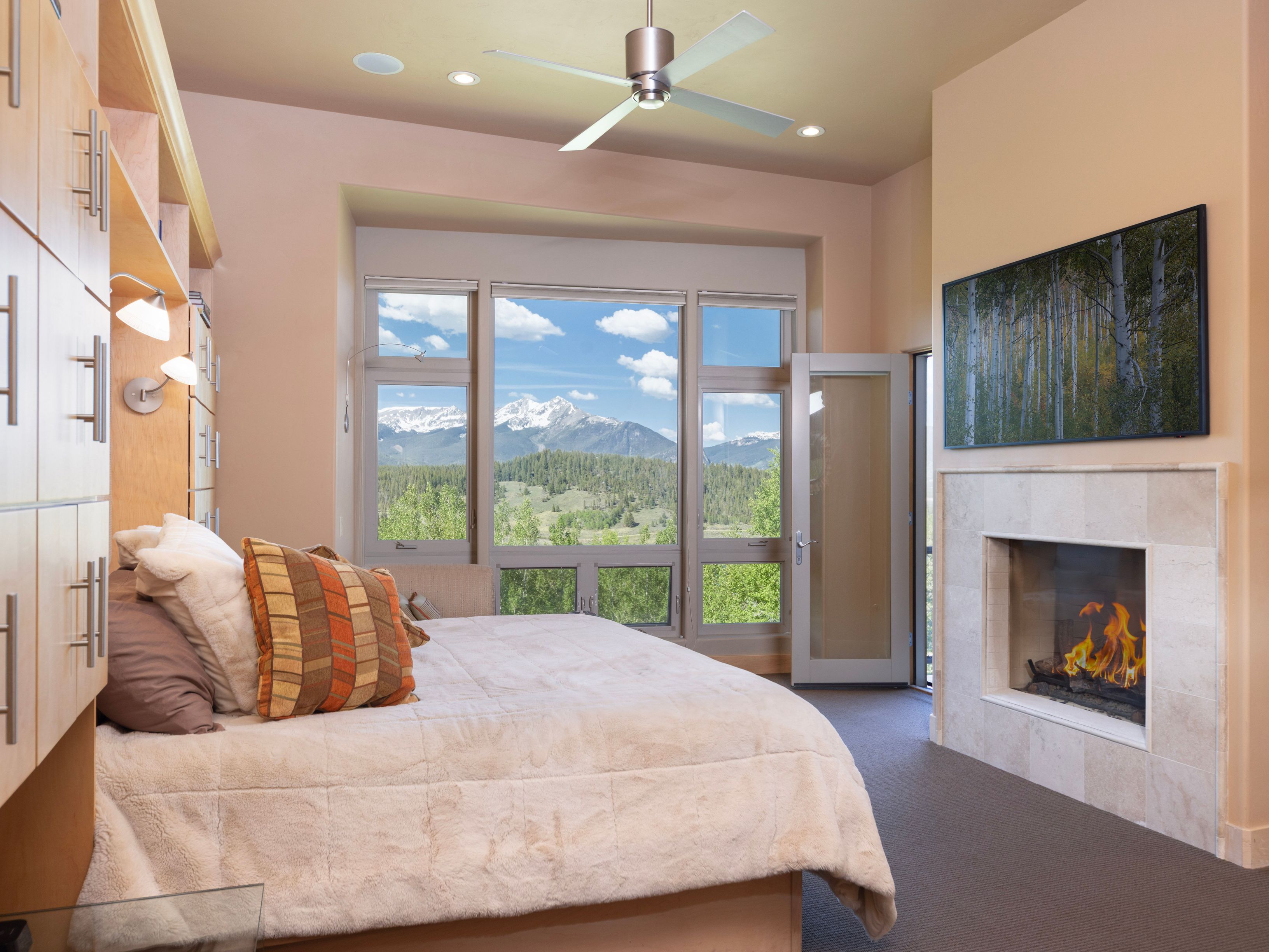 This primary bedroom features a cozy, neutral-toned aesthetic with a large bed positioned to face a wall-mounted television and a stone-clad fireplace. To the left, light wood cabinetry provides ample storage, while a set of large windows offers a stunning, unobstructed view of the mountain landscape. A glass door adjacent to the fireplace provides direct access to the outdoors, creating a seamless connection between the interior space and the natural surroundings.