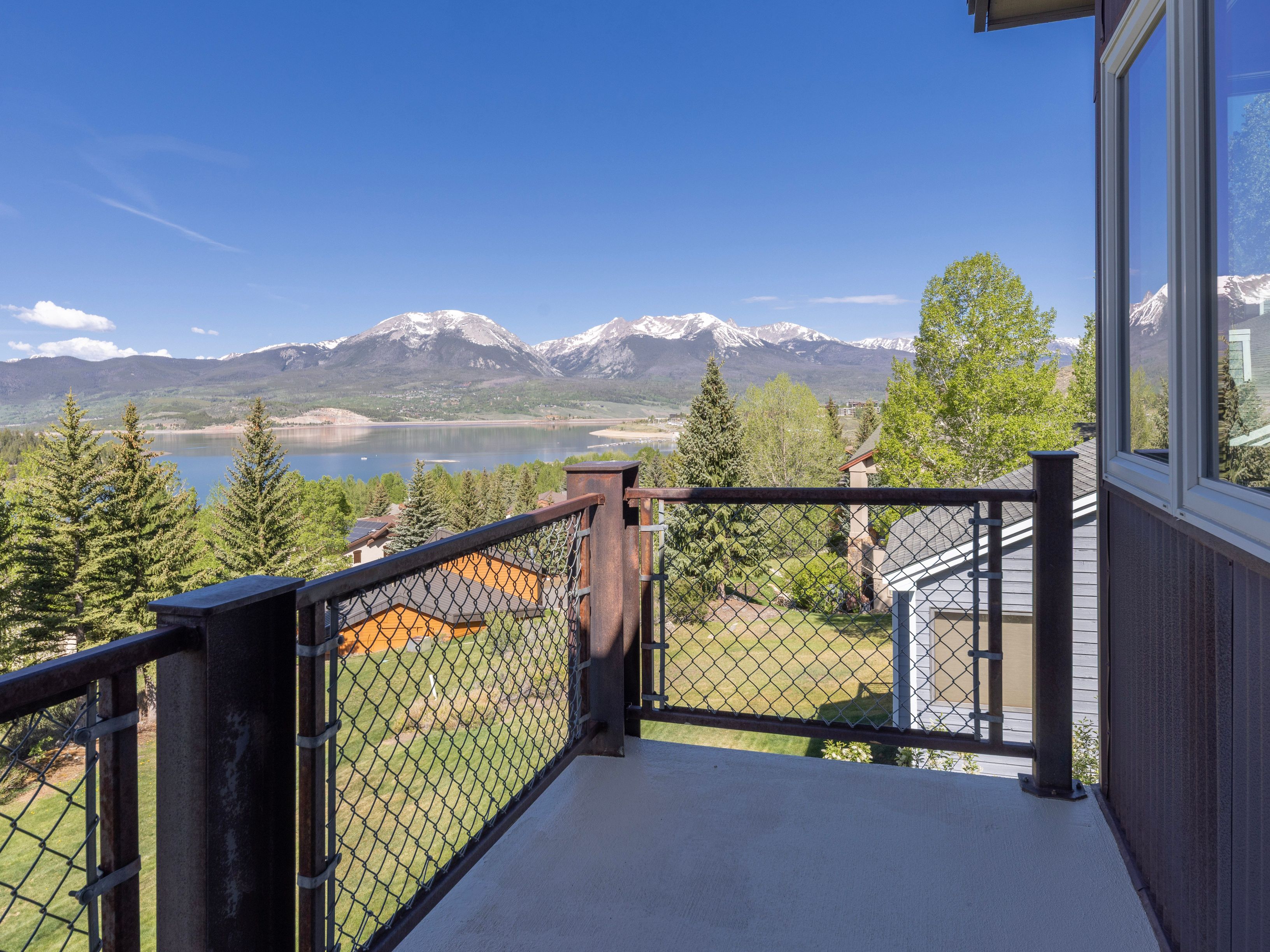 This elevated balcony offers a stunning, panoramic view of a serene lake framed by majestic, snow-capped mountain peaks under a clear blue sky. The deck features a sturdy, industrial-style railing with metal posts and chain-link mesh, providing a secure yet open vantage point. The perspective captures the transition from the private outdoor living space to the expansive natural landscape, highlighting the property's prime location.