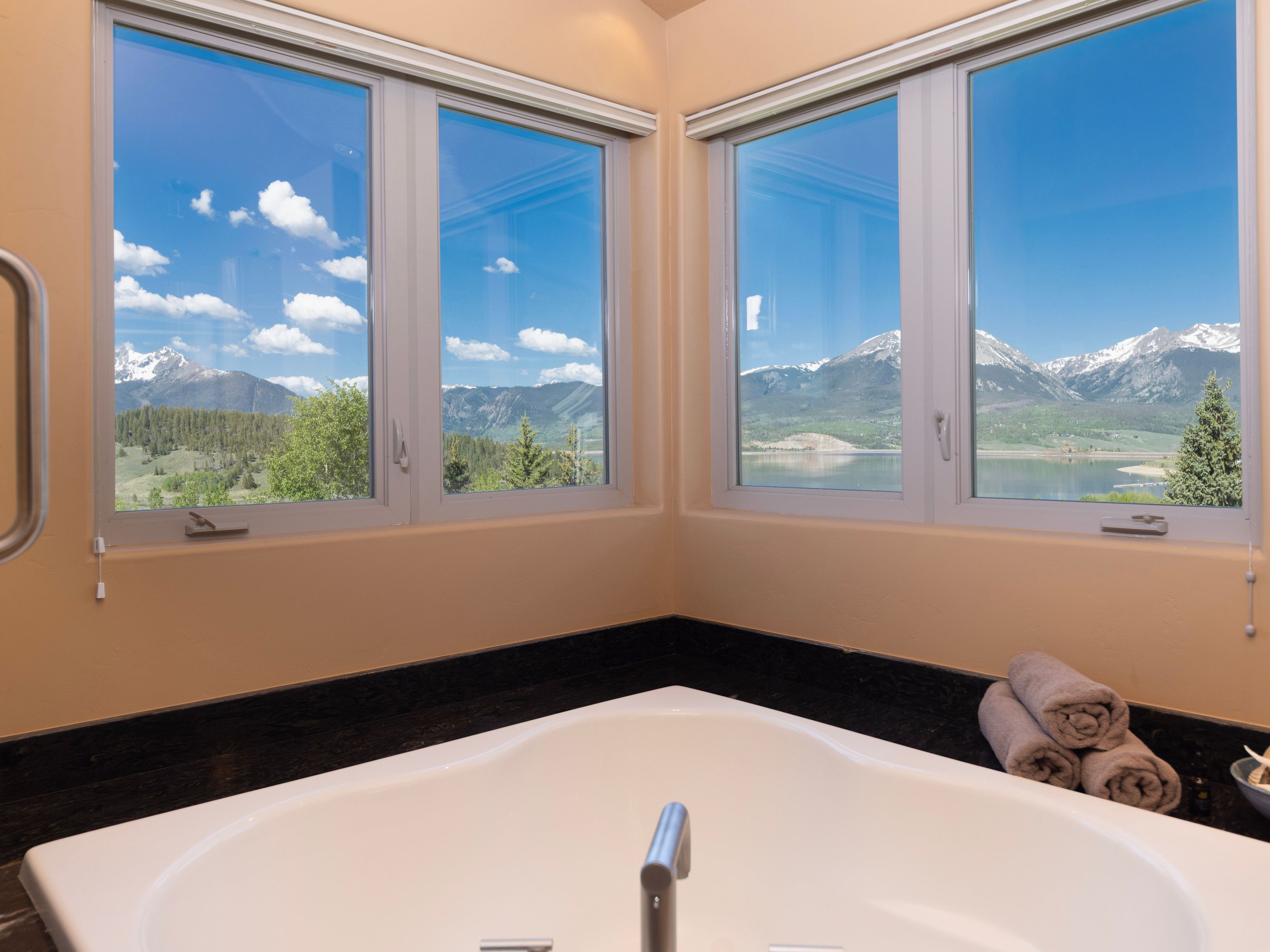This primary bathroom features a corner soaking tub set against a dark stone deck, positioned perfectly to take advantage of the expansive mountain and lake views through large windows. The neutral-toned walls provide a calm backdrop, while a stack of soft towels rests on the corner ledge, adding a touch of spa-like comfort. The perspective is centered on the tub, emphasizing the serene connection between the indoor space and the natural landscape outside.