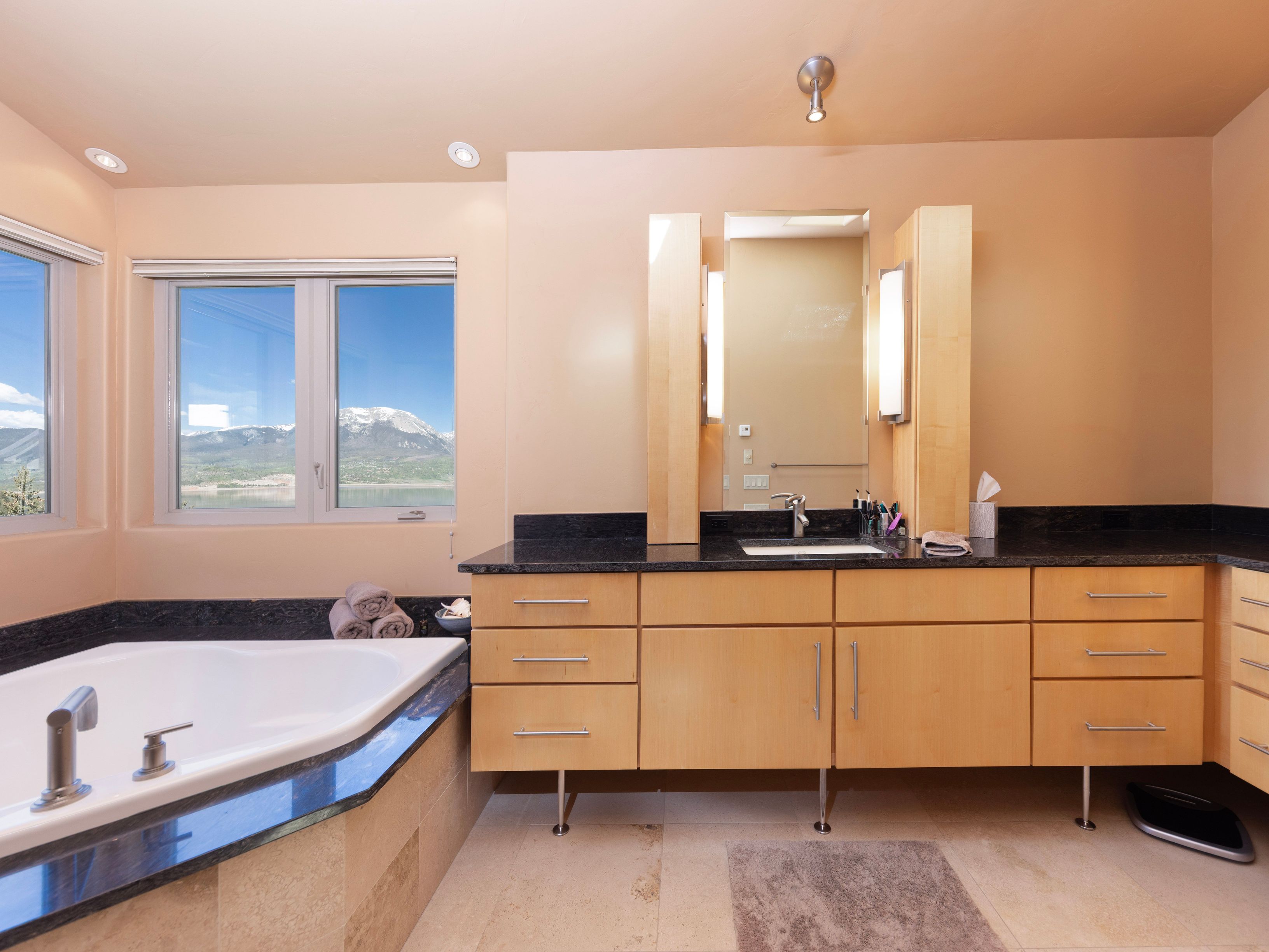 This primary bathroom features a light wood vanity with a dark stone countertop and a large mirror flanked by vertical light fixtures. To the left, a corner soaking tub is positioned beneath large windows that offer a scenic view of a lake and snow-capped mountains. The space is finished with neutral-toned tile flooring and a minimalist aesthetic, creating a bright and serene atmosphere.