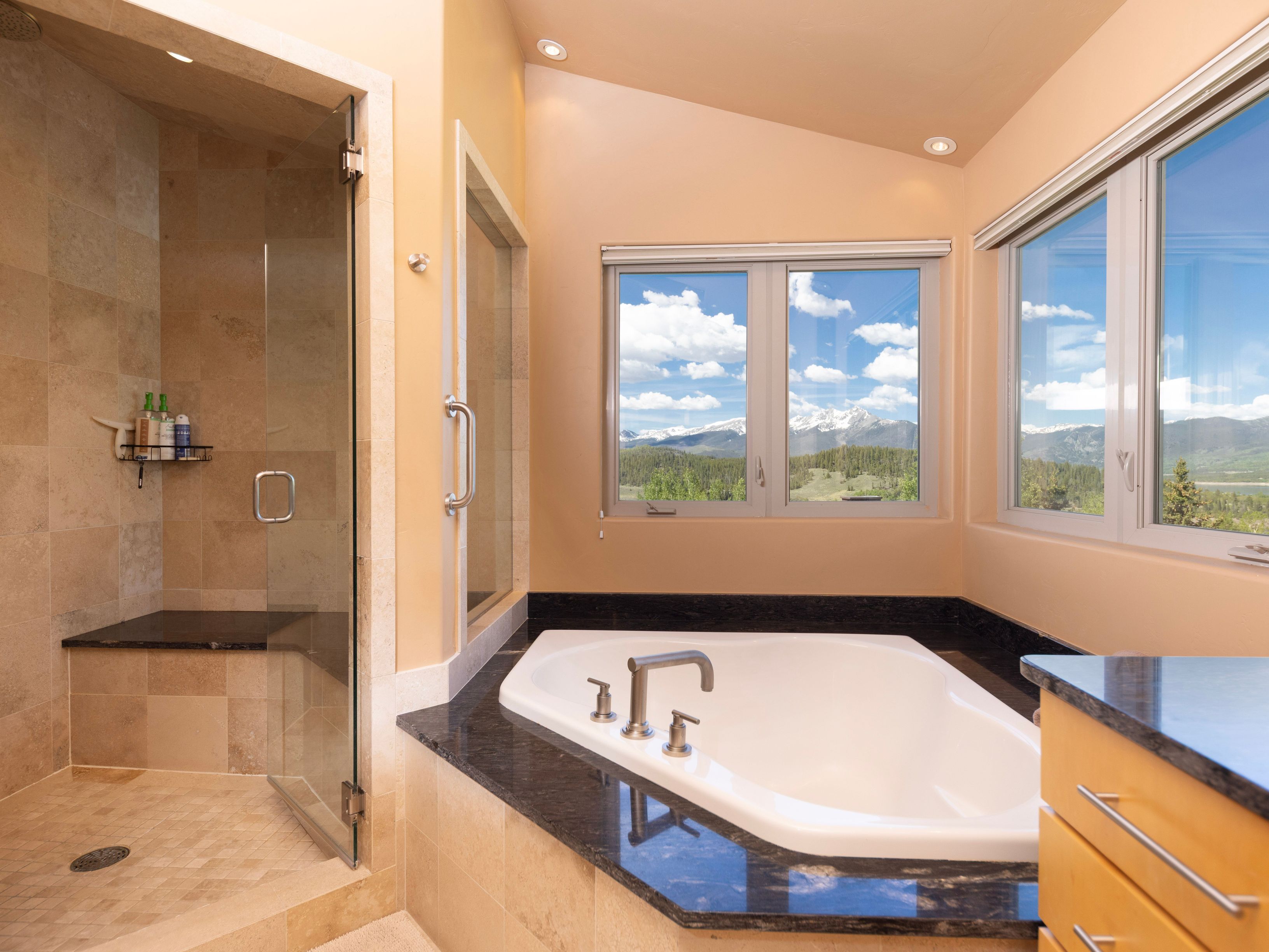 This primary bathroom features a corner soaking tub set into a dark stone deck, positioned perfectly to take advantage of the expansive mountain views through large windows. To the left, a glass-enclosed walk-in shower with stone tile walls and a built-in bench provides a spa-like experience. The room is finished with light-toned walls and light wood cabinetry, creating a bright and serene atmosphere.