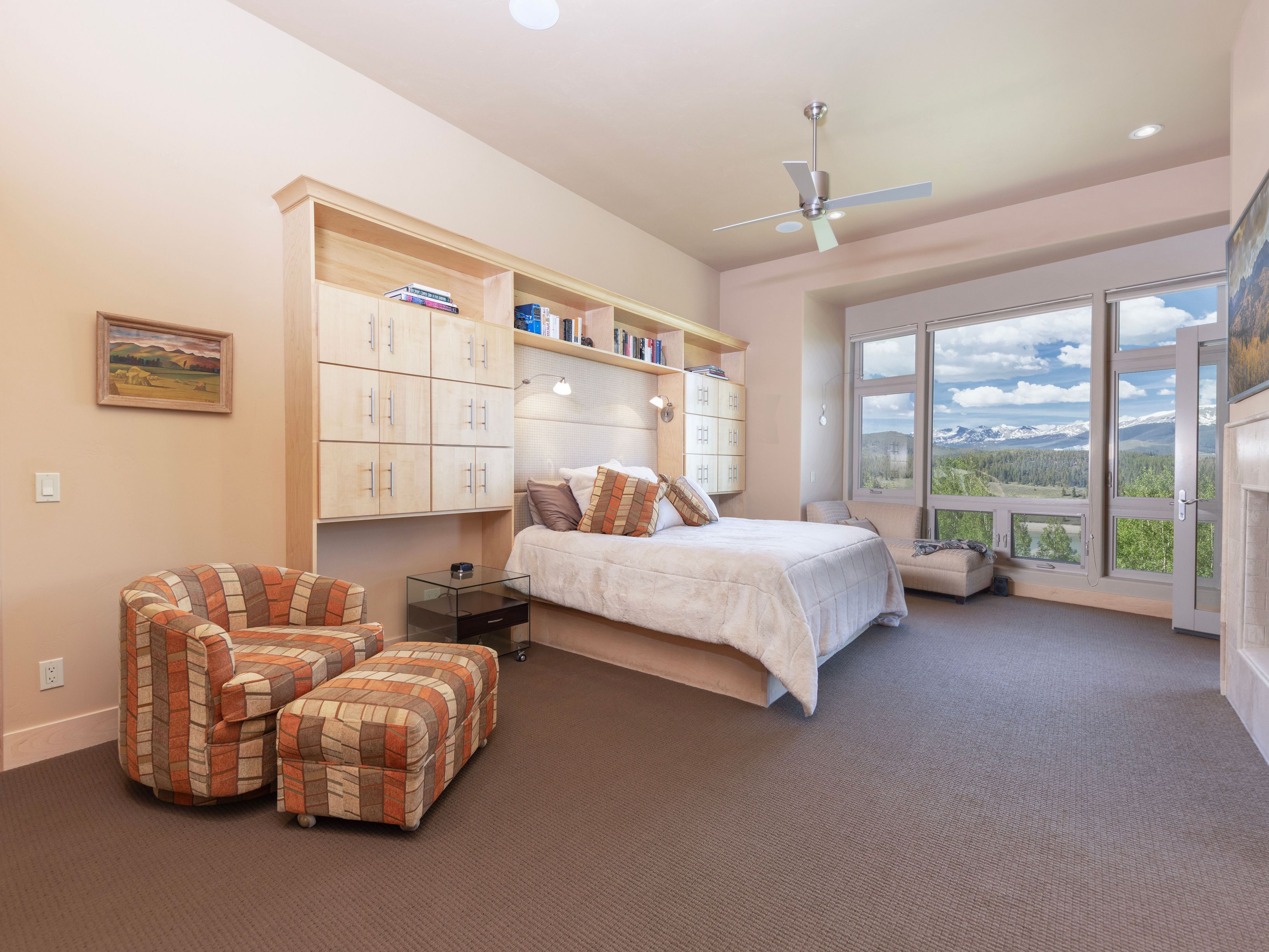 This spacious primary bedroom features a light wood built-in storage unit surrounding the headboard, complemented by a comfortable armchair and ottoman in a warm, patterned fabric. Large windows provide an expansive view of the surrounding mountains and landscape, filling the room with natural light. The neutral color palette and clean lines create a serene and inviting atmosphere.